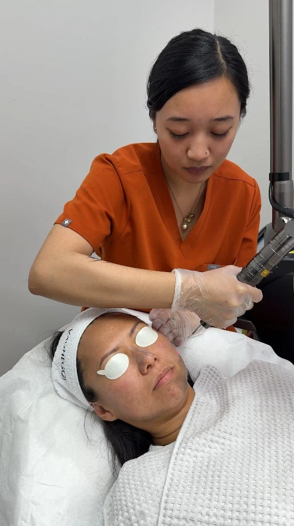 A woman receiving a cosmetic treatment with a handheld device from a professional in an orange uniform in a clinic setting.