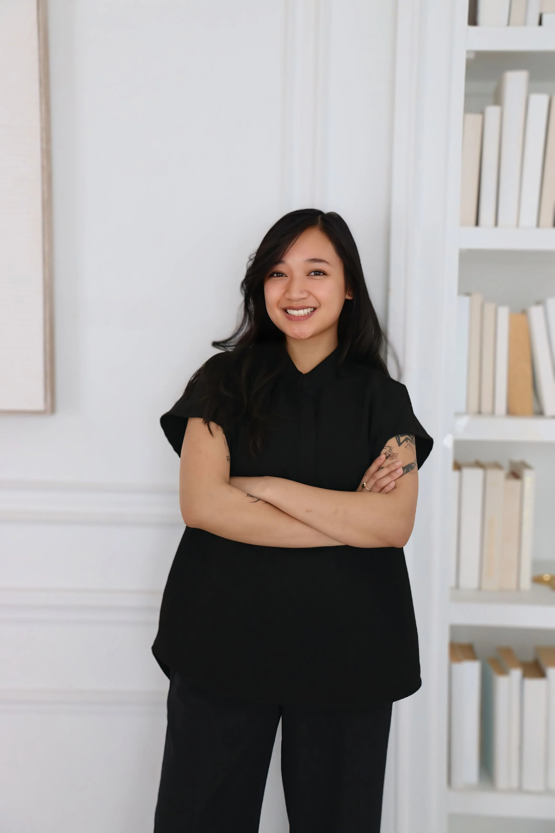 A woman with dark shoulder-length hair, wearing a white sleeveless vest and beige pants, sitting on a light-colored sofa in a minimalist room with neutral tones, decorative objects on shelves, and artwork on the wall.