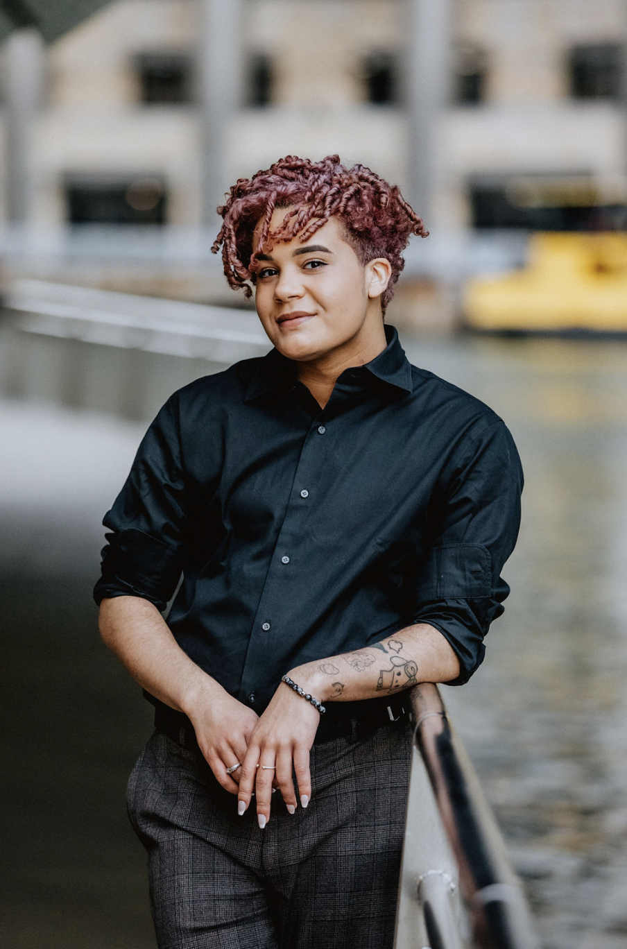 Portrait of a person with curly red hair in a dark button up shirt near a body of water.