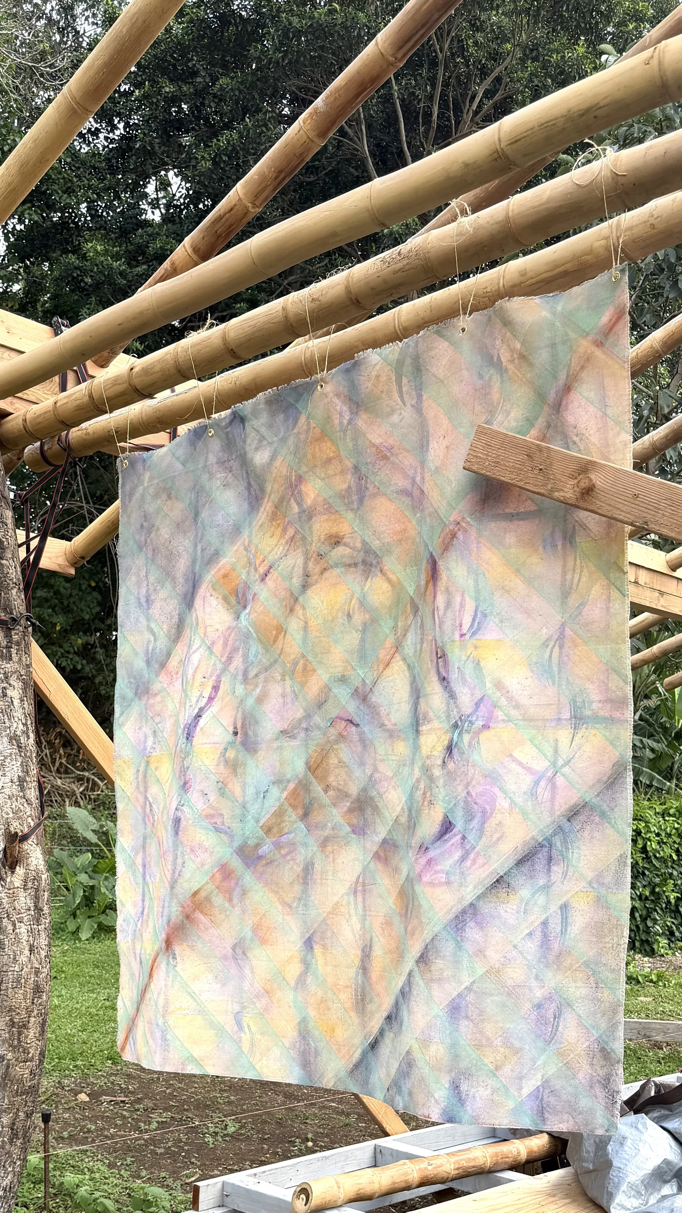 Colorful fabric with abstract pattern hanging on bamboo rods, outdoors in a garden setting.