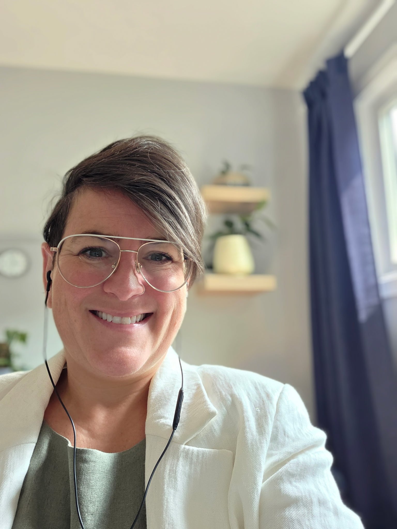 A woman with short, brown hair and glasses smiling at the camera, wearing a white blazer and a light gray shirt, with a window with blue curtains and a decorative shelf with a lamp in the background.