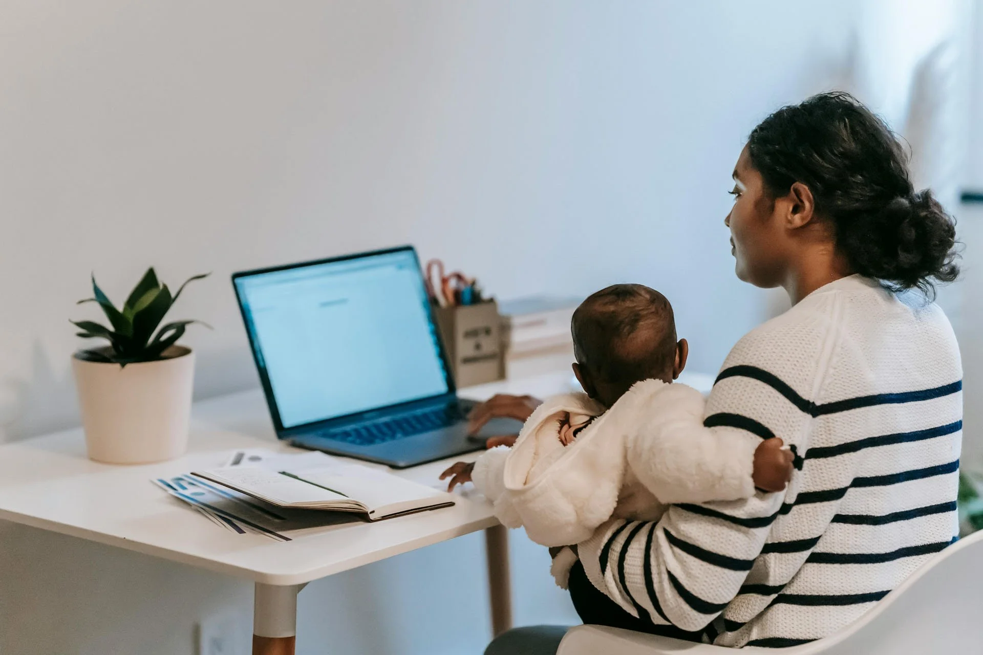 A woman holds a baby while sitting at a desk with a laptop, a potted plant, and paperwork in a bright room.