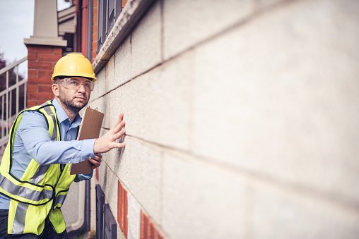  Building inspector, architect, engineer, general contractor, repairman insurance adjuster, or other blue collar worker inspecting a residential building. Wearing a safety vest, yellow helmet and clear safety glasses, he is checking very meticulously