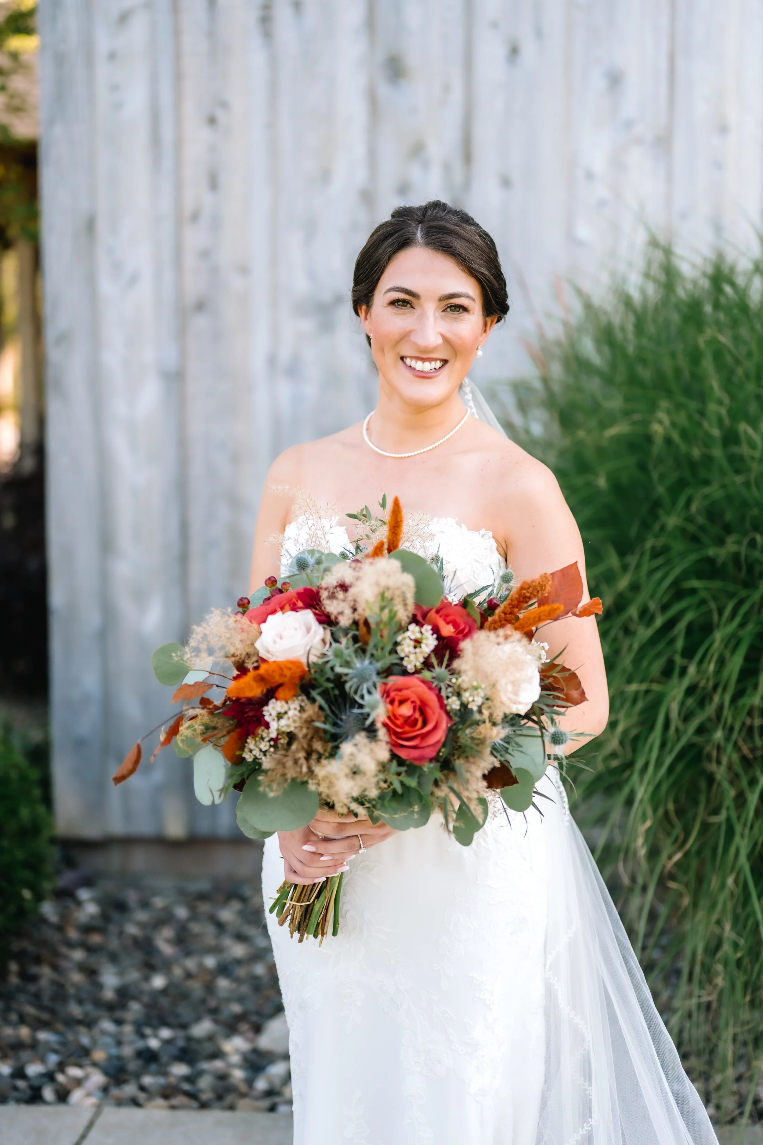 A bride in a white wedding dress holding a bouquet of flowers standing outdoors with a wooden fence and green plants in the background.