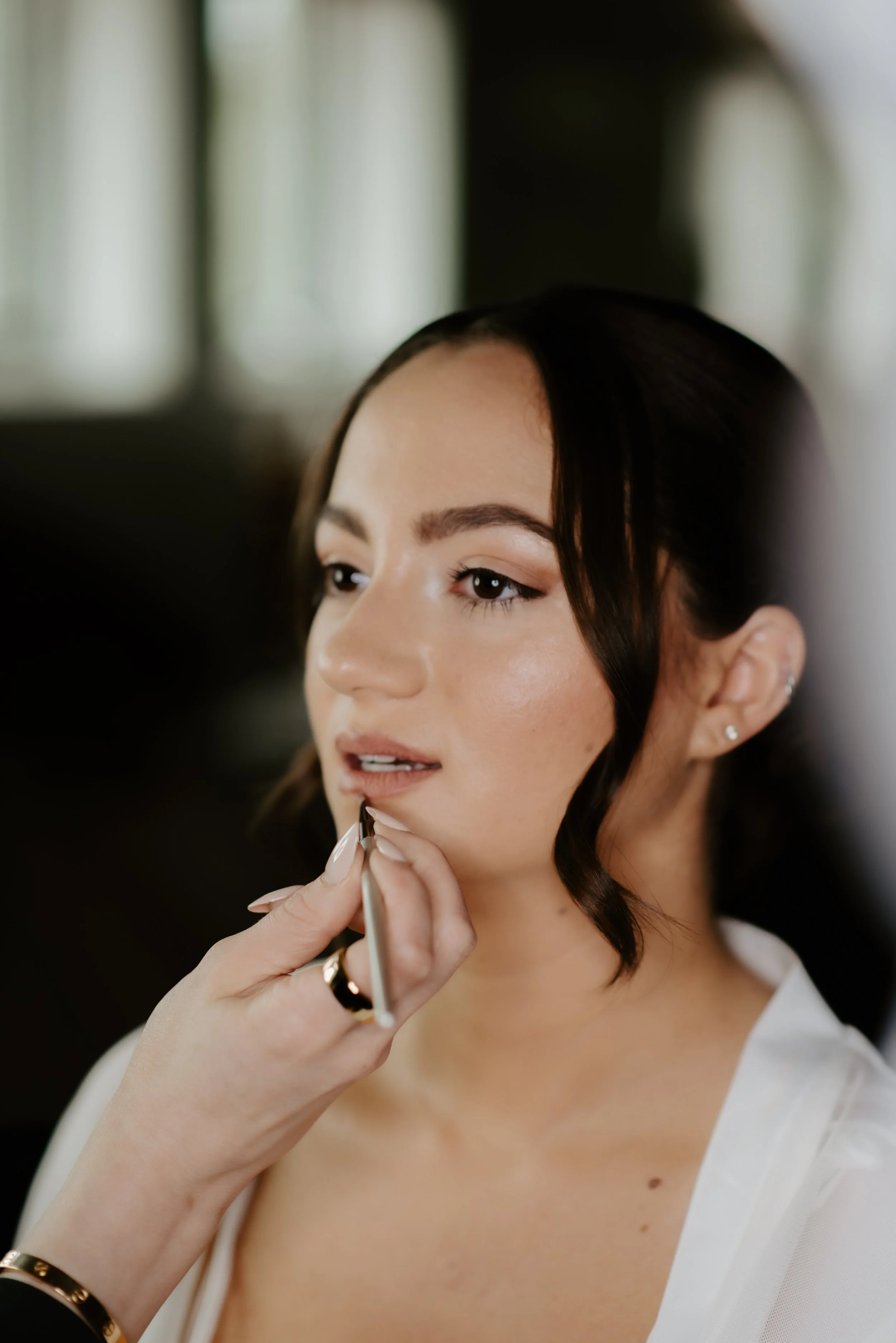 A woman with dark hair and earrings having makeup applied to her lips by a makeup artist in a well-lit room.