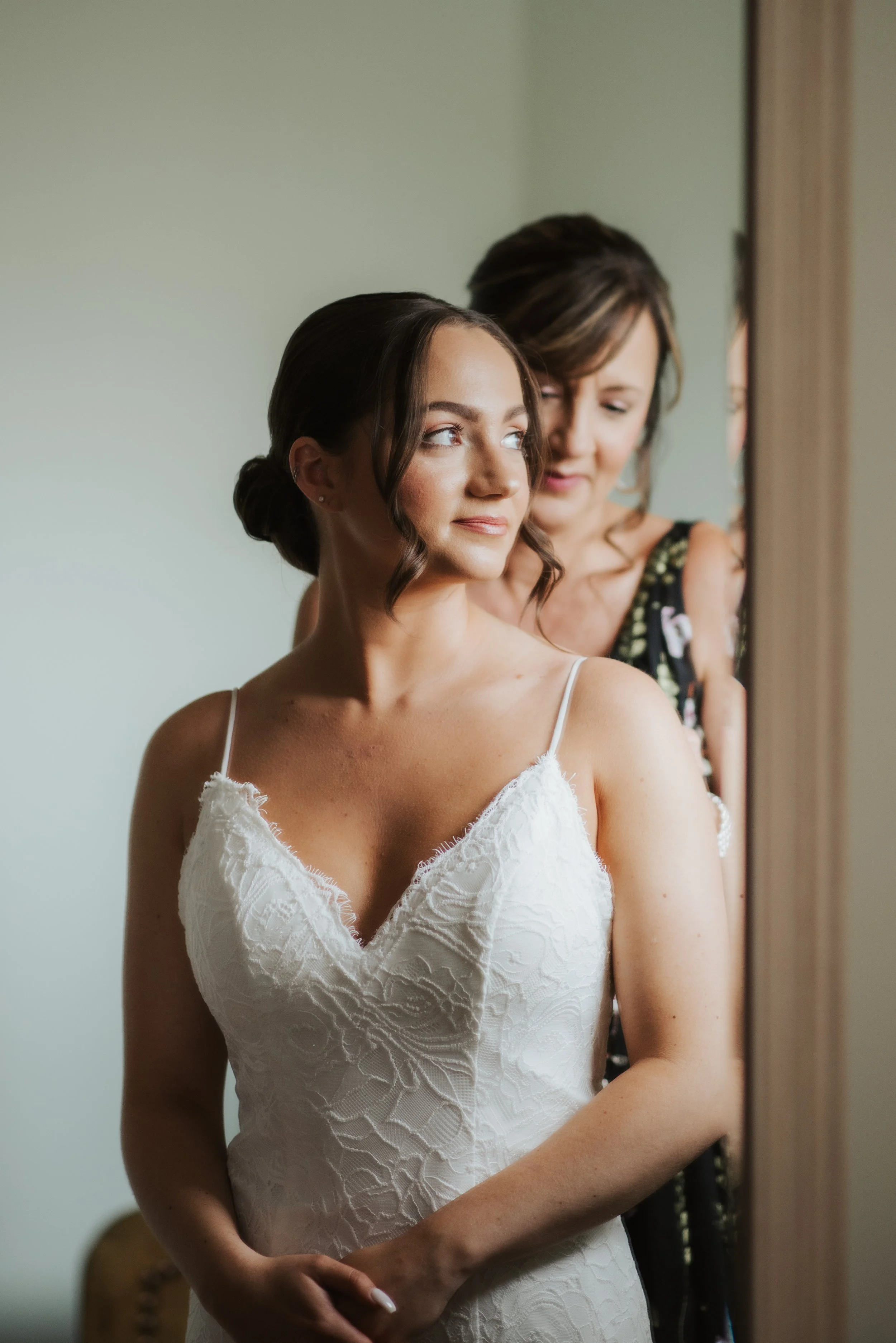 A bride in a white lace dress looks into a mirror while a woman behind her adjusts her hair or dress.