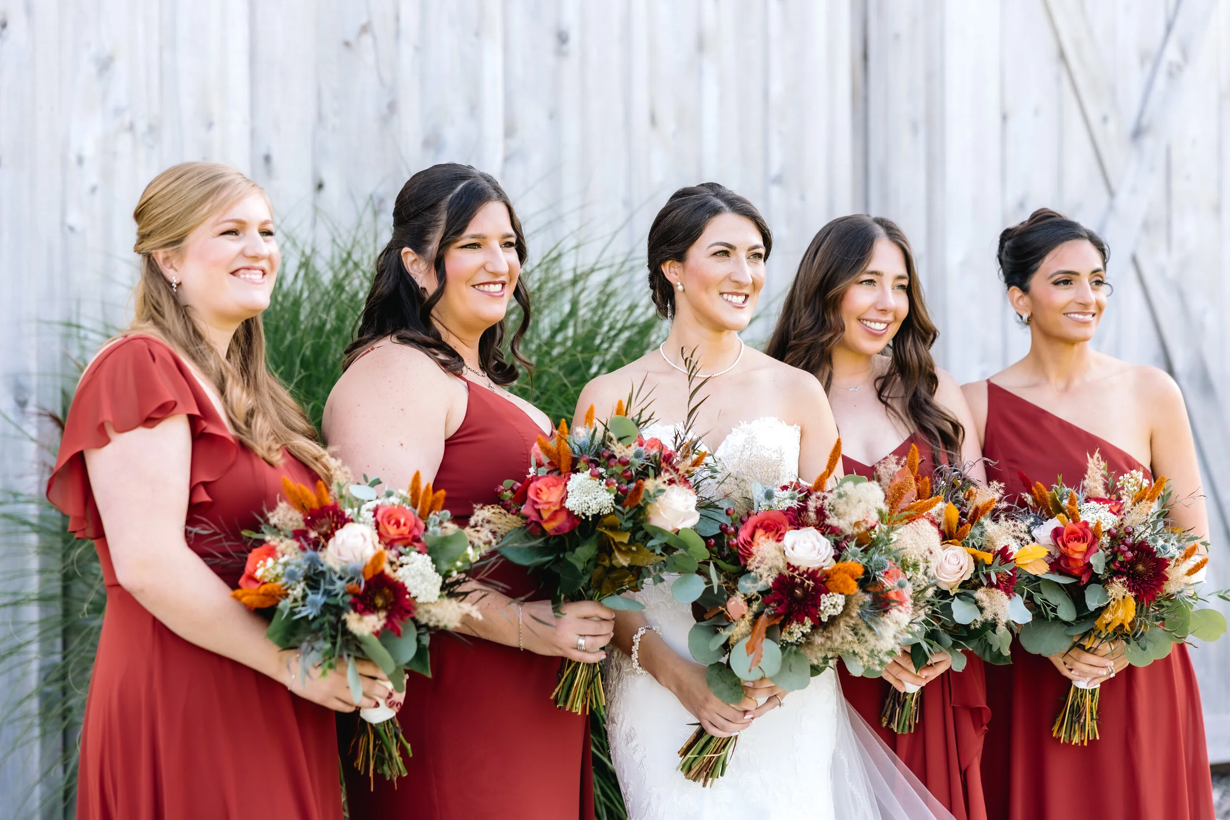 Bride in white wedding dress and five bridesmaids in red dresses standing outdoors, each holding a colorful bouquet of flowers, smiling.