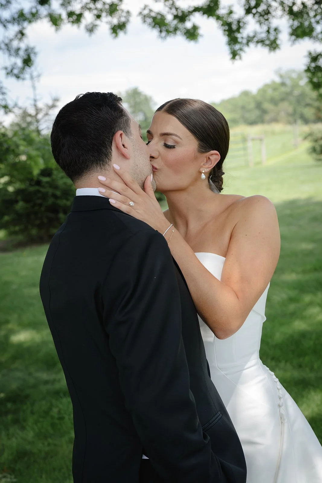 A bride and groom share a kiss outdoors on a sunny day, with trees and greenery in the background.