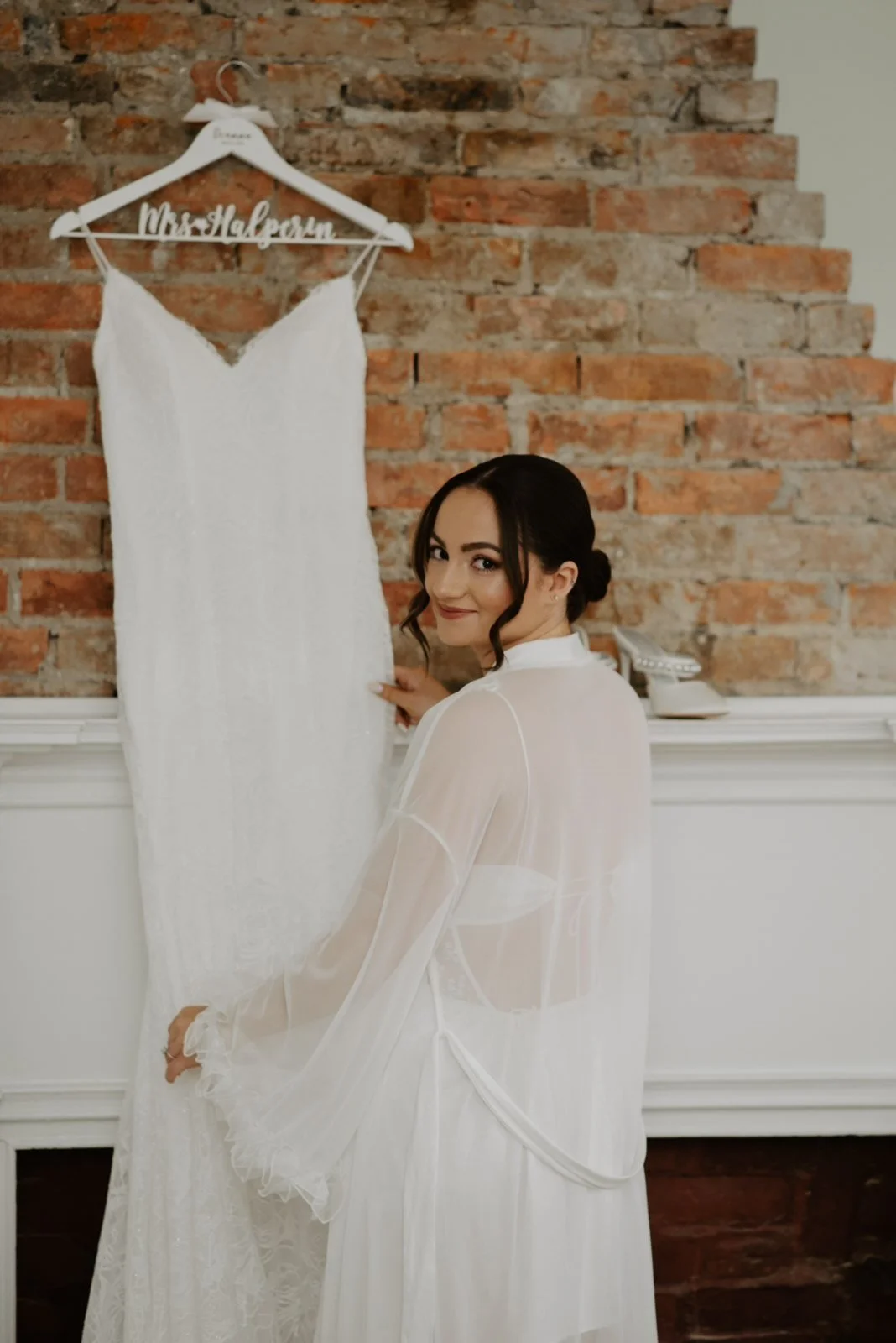 A bride with dark hair styled in an updo, wearing a sheer white robe, stands next to a white wedding dress hanging on a brick wall. The dress has a hanger with a custom name tag reading 'Mrs. Halevson'.