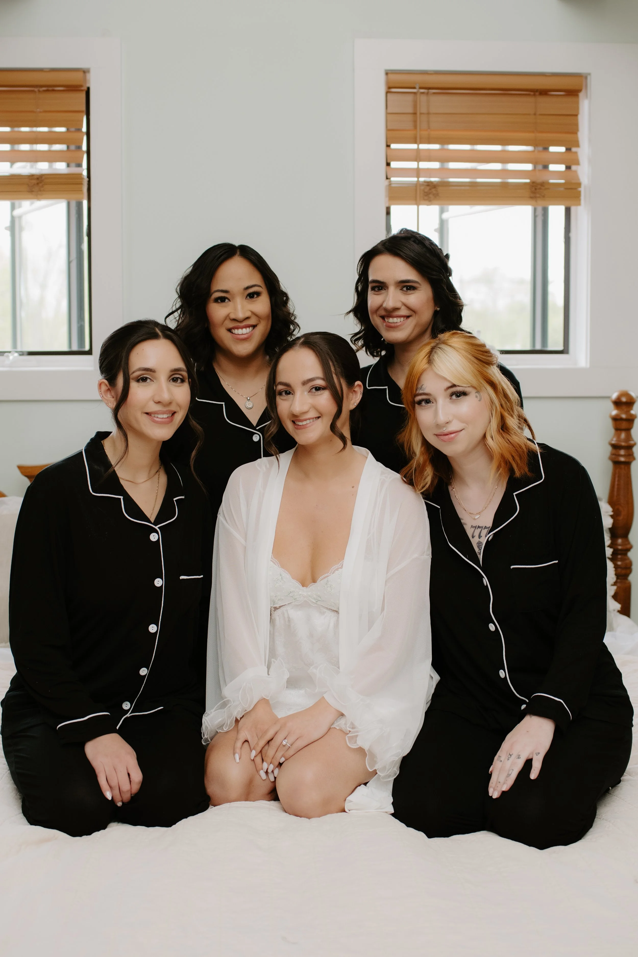 A bride in white lingerie and a sheer robe sitting on a bed surrounded by five women wearing matching black pajamas with white piping in a bedroom with two windows and wooden blinds.