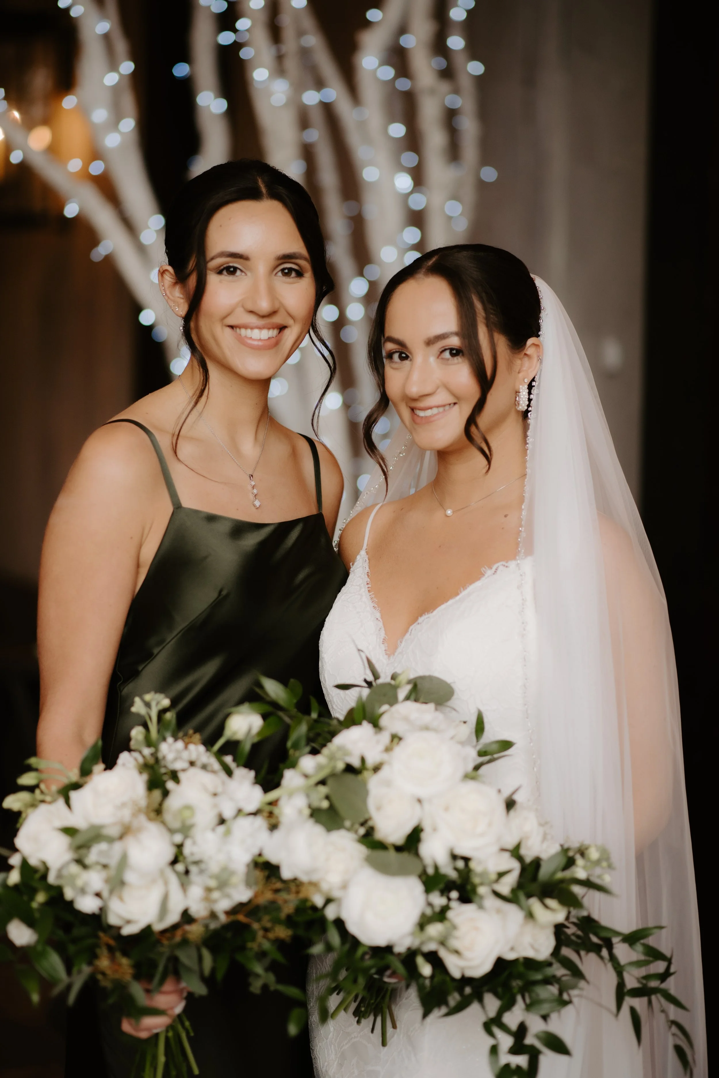 Two women, one in a black dress and the other in a wedding gown with a veil, smiling and holding a large bouquet of white flowers, with string lights in the background.