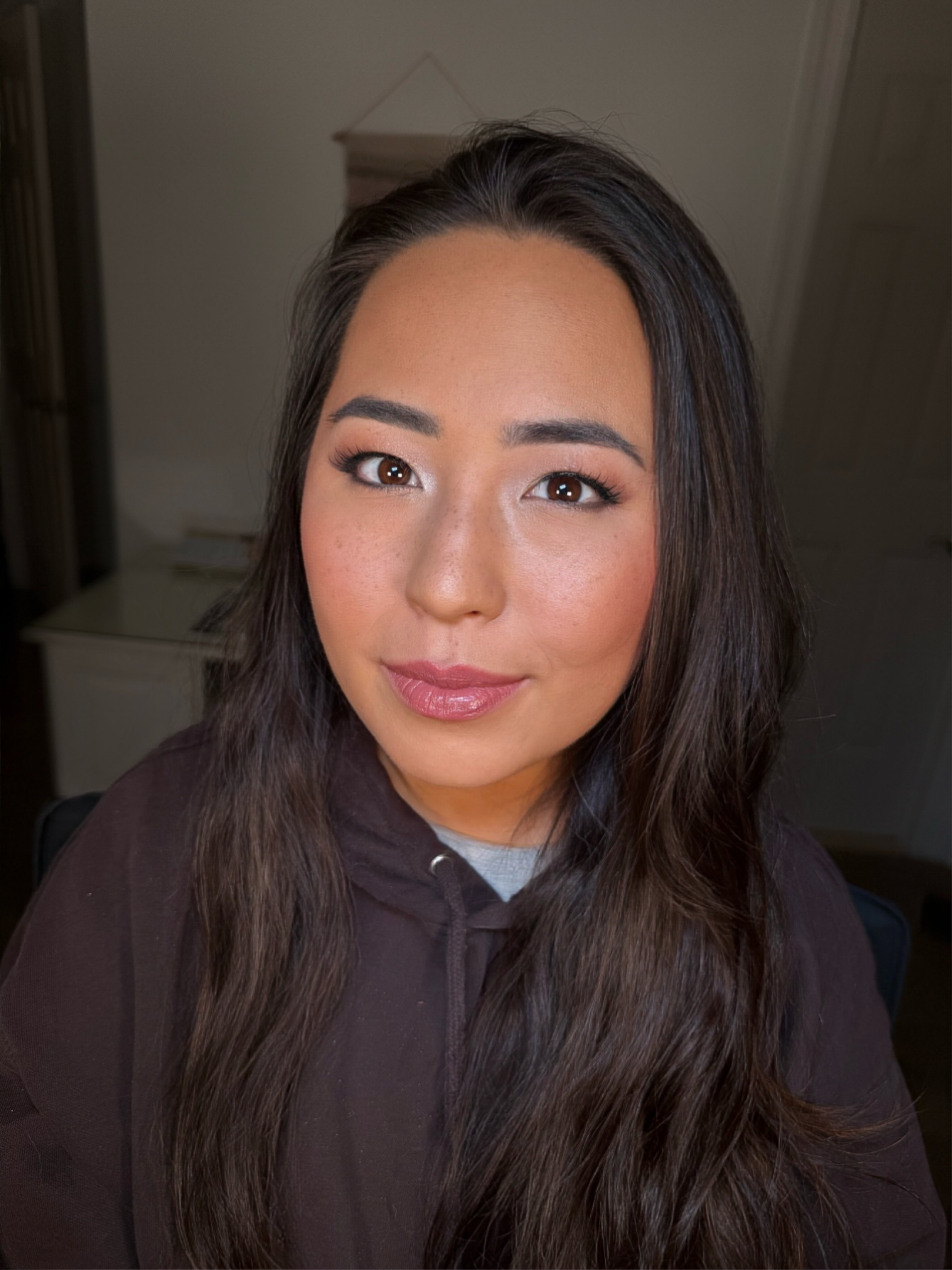 Close-up of a woman with long dark hair, smiling, wearing a dark hoodie, in a room with a white wall and a decorative hanging in the background.