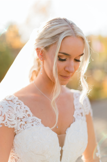 A bride with blonde hair styled in a loose updo, wearing a lace wedding dress and a veil, standing outdoors with sunlight in the background.