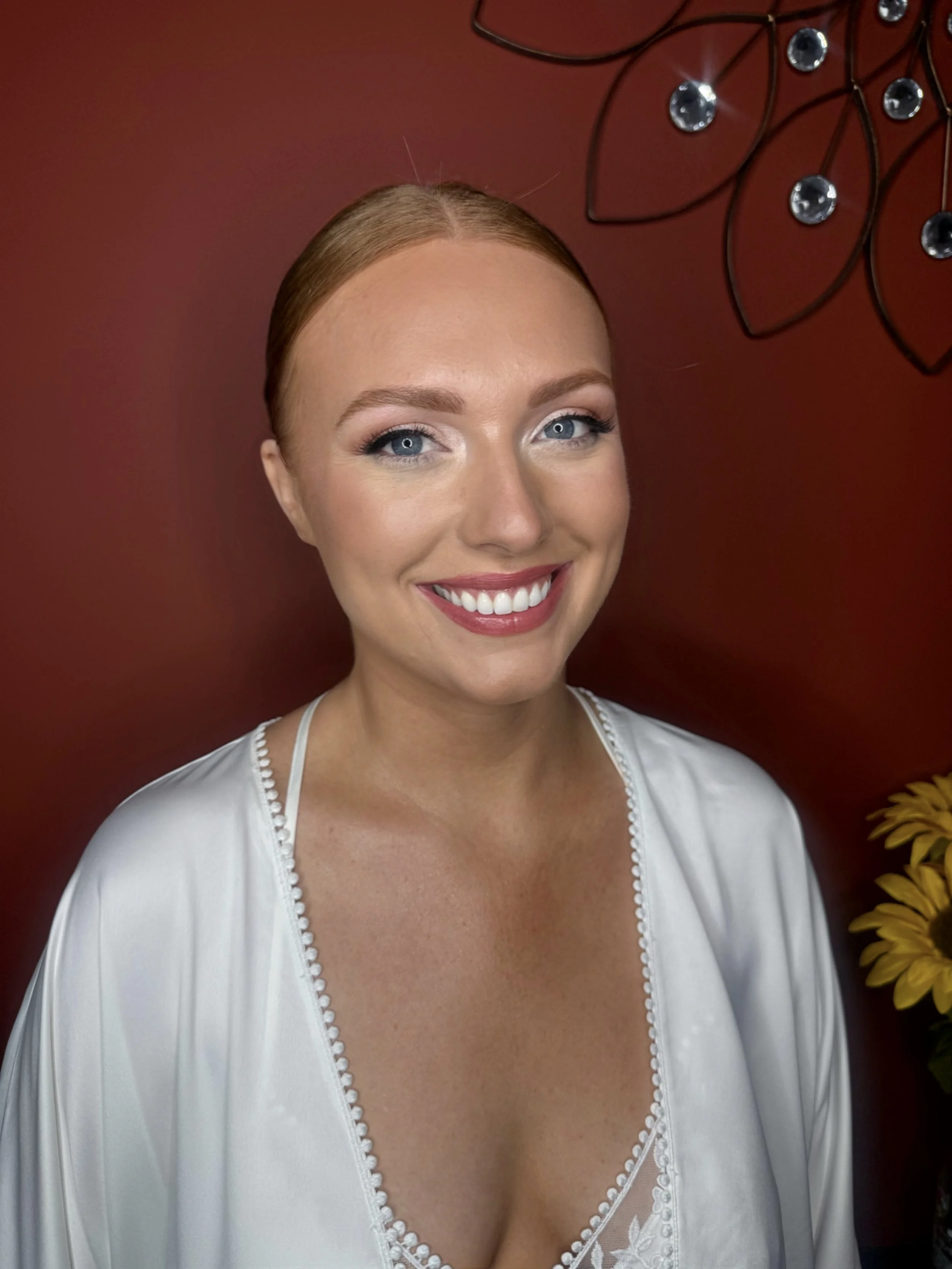 A smiling woman with red hair in a bun, wearing a white satin robe with lace trim, standing in front of a red wall with decorative metal art and a vase of yellow flowers.