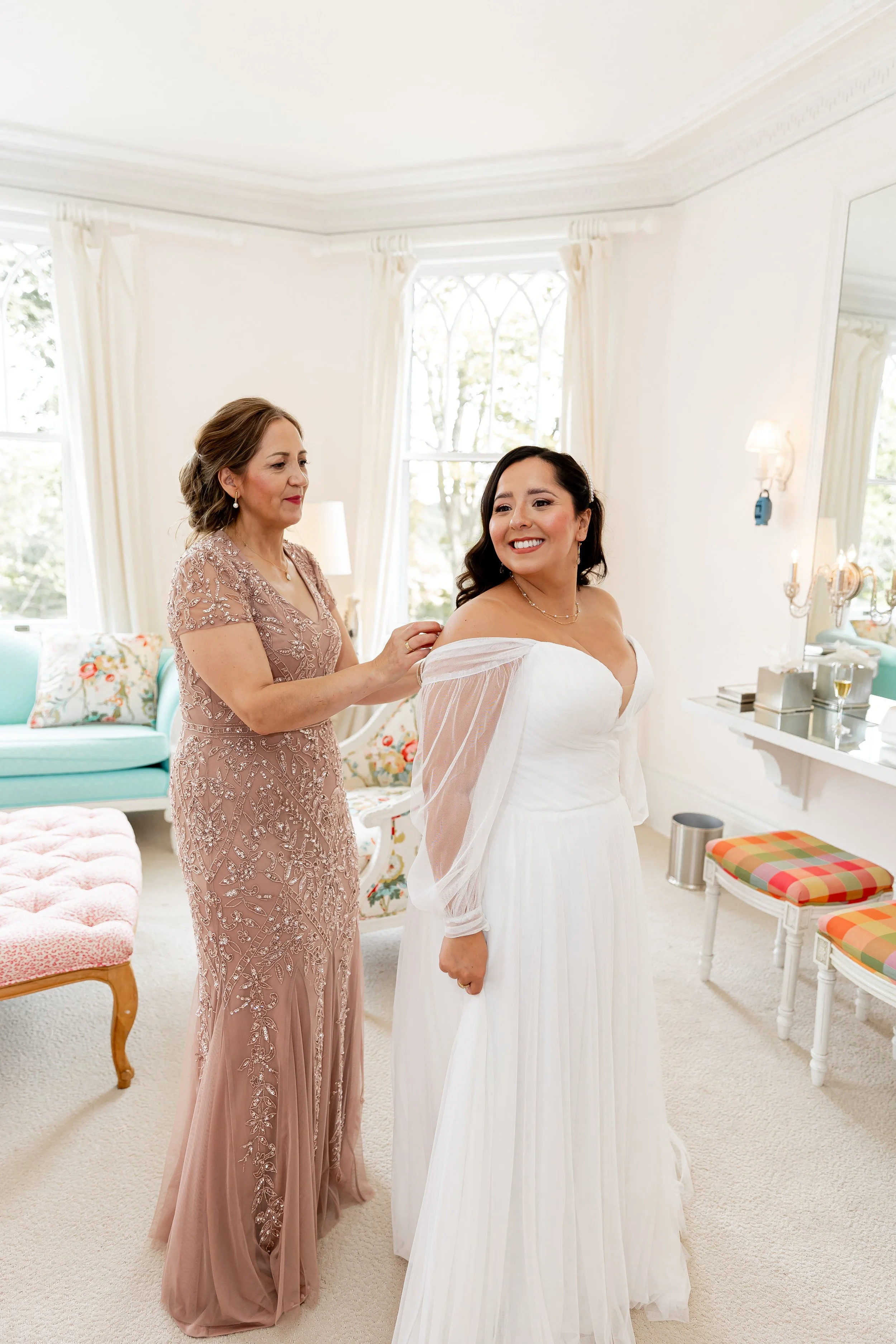 A bride in a white dress and a woman in a pink dress are in a bright, elegant room, she is helping the bride with her dress.