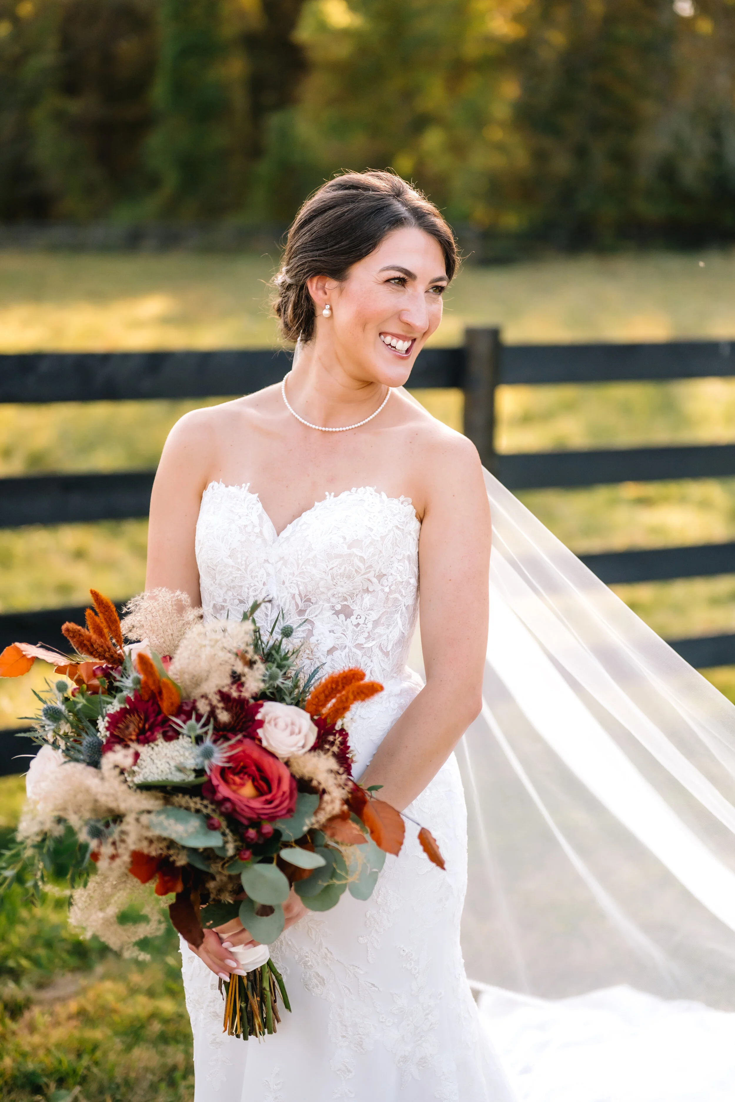 A bride in a white lace wedding dress holding a bouquet of colorful flowers, smiling outdoors with a black wooden fence and trees in the background.