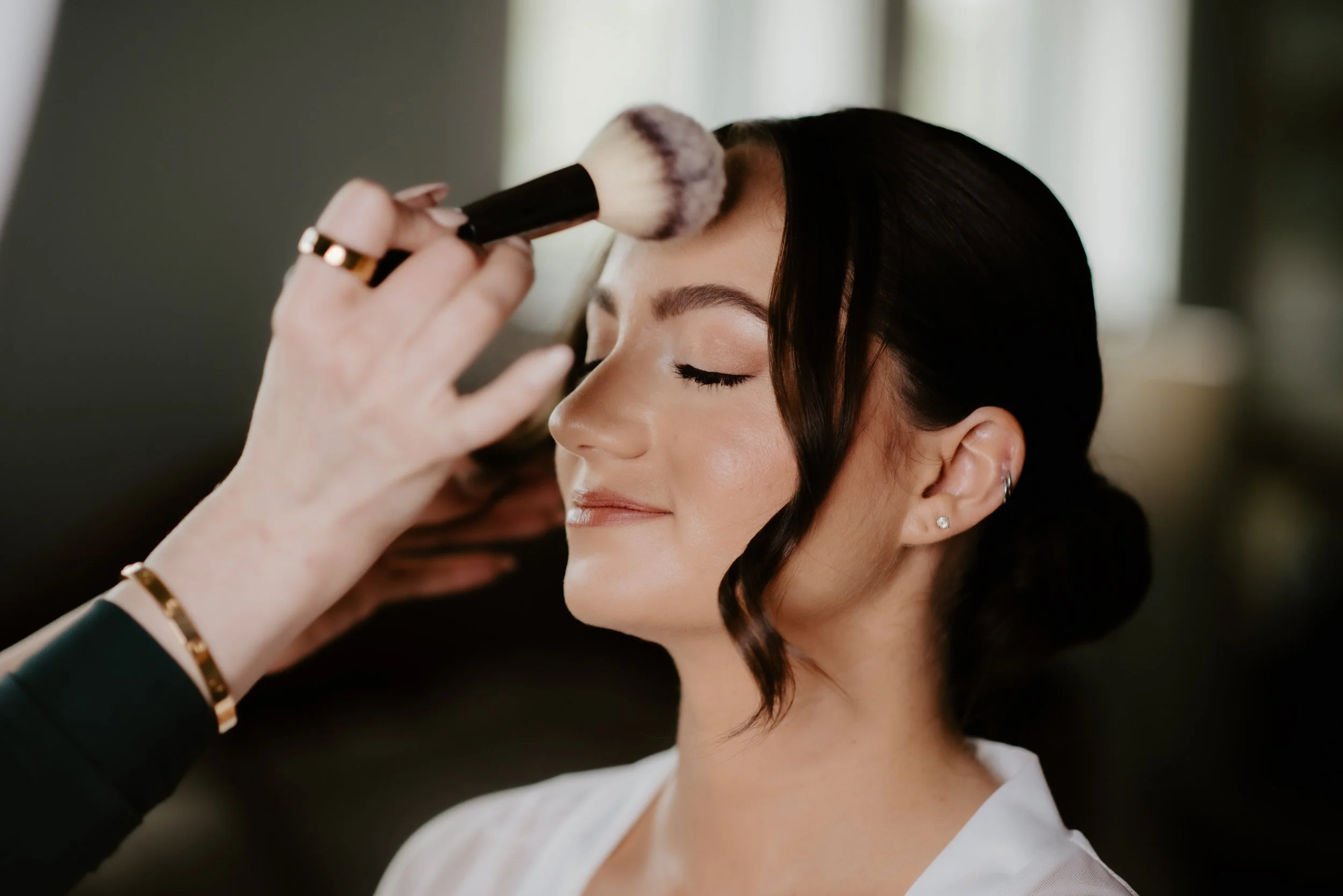 Close-up of a woman with dark hair getting her makeup applied, her eyes closed, by a makeup artist holding a large fluffy brush. The woman is smiling softly.