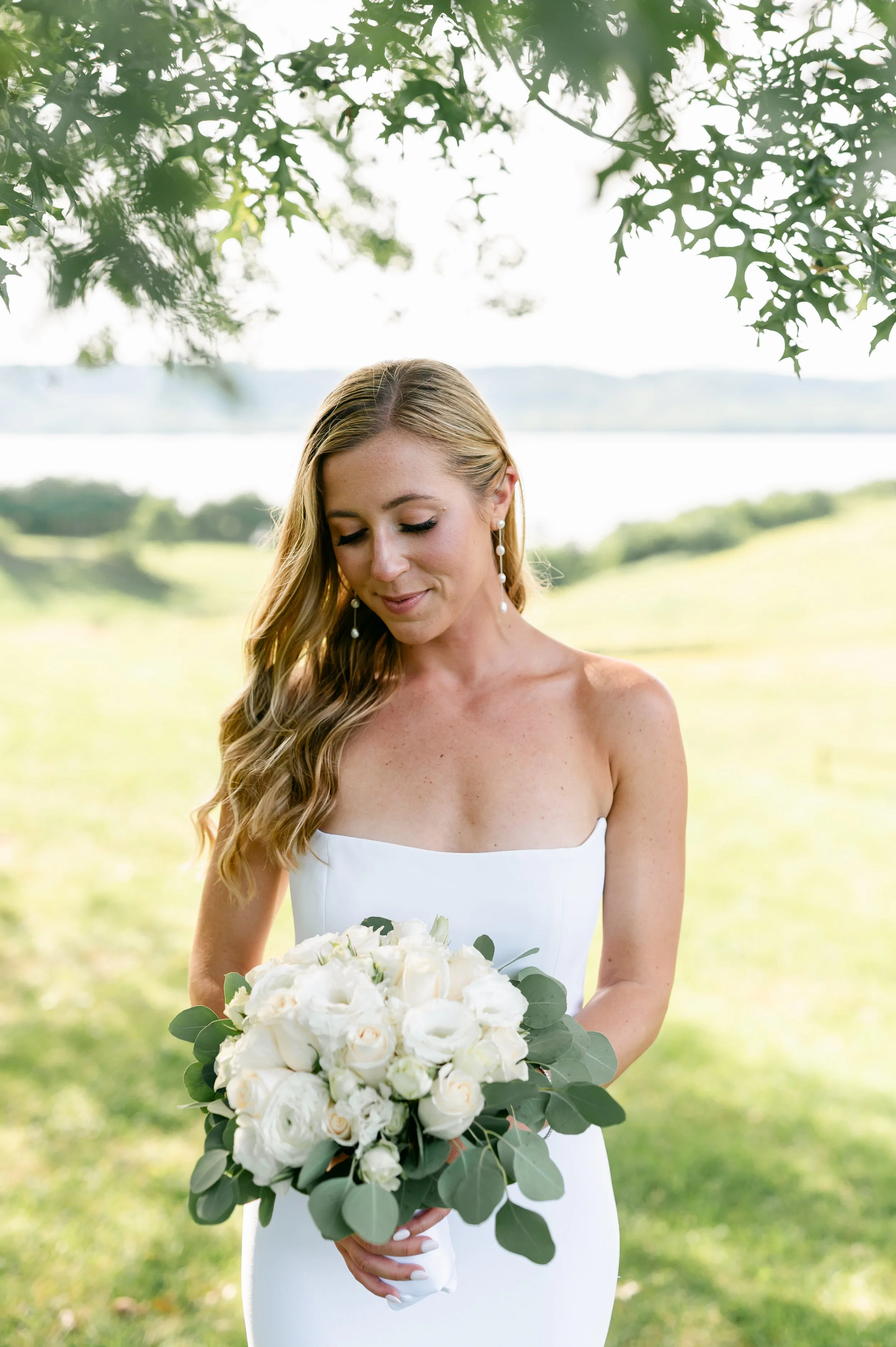 A bride holding a bouquet of white roses and greenery, standing outdoors in a green landscape with a body of water in the background.