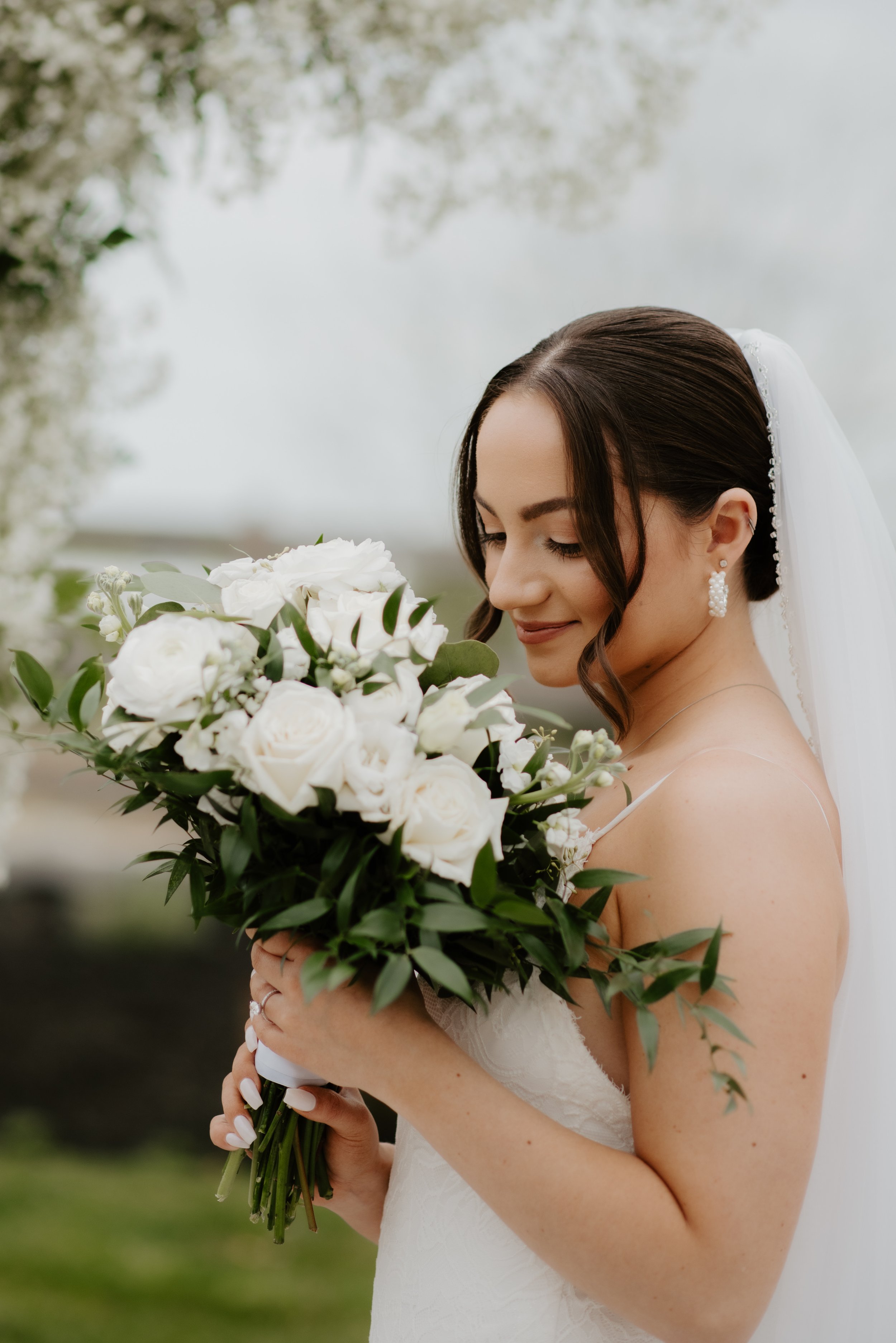 A bride with dark hair styled in soft waves, wearing drop earrings, a white wedding dress, and a veil, holding a large bouquet of white roses and greenery outdoors.