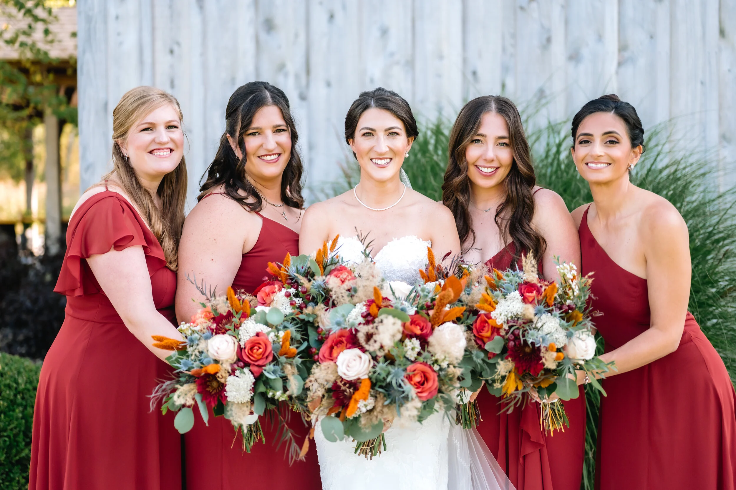 A bride in a white wedding dress is standing with four bridesmaids in red dresses, all holding large floral bouquets, outdoors near a wooden fence.