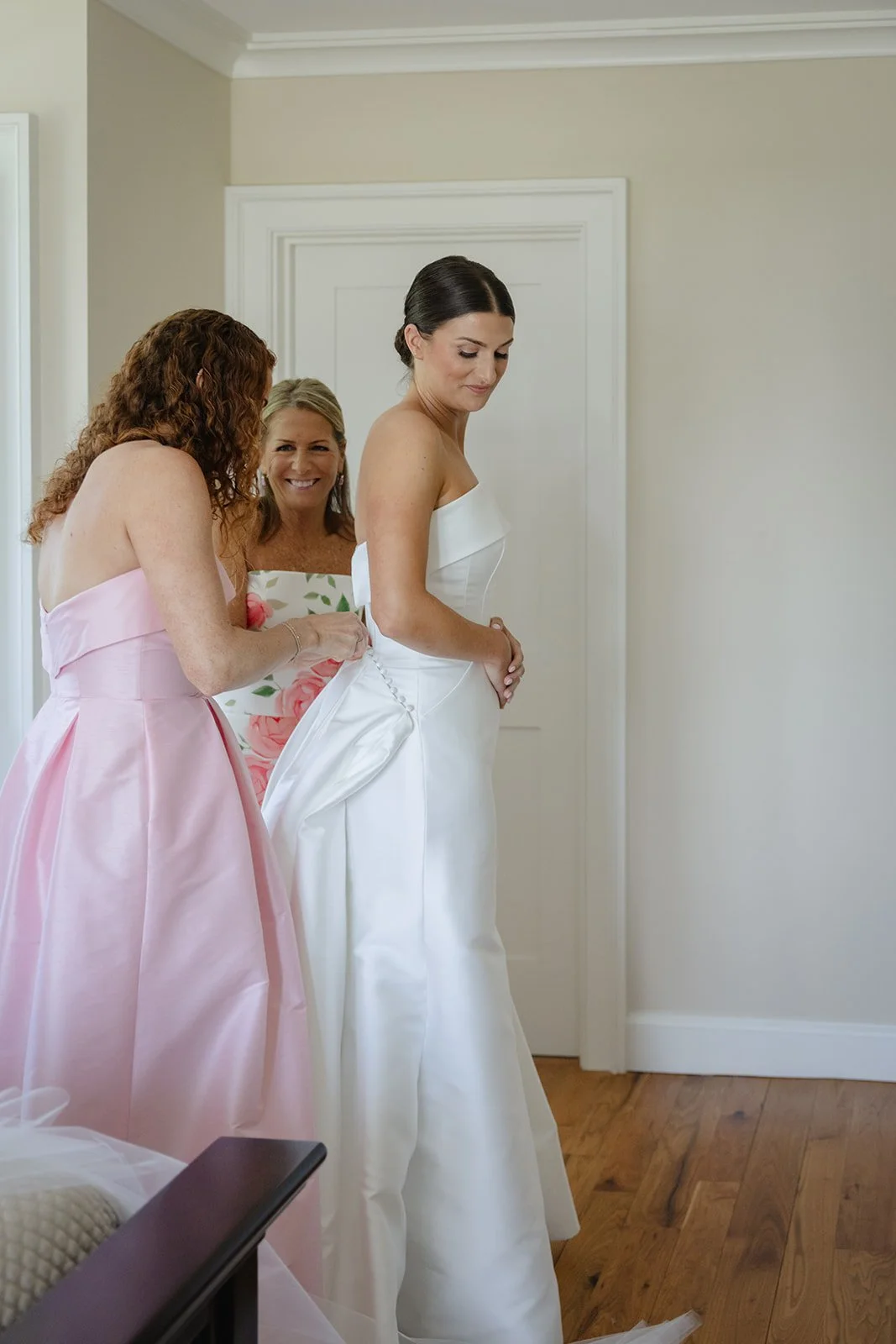 A bride in a white wedding dress stands while two women, one in a pink dress and the other in a floral dress, help her with her dress, and they all smile during a wedding preparation scene.