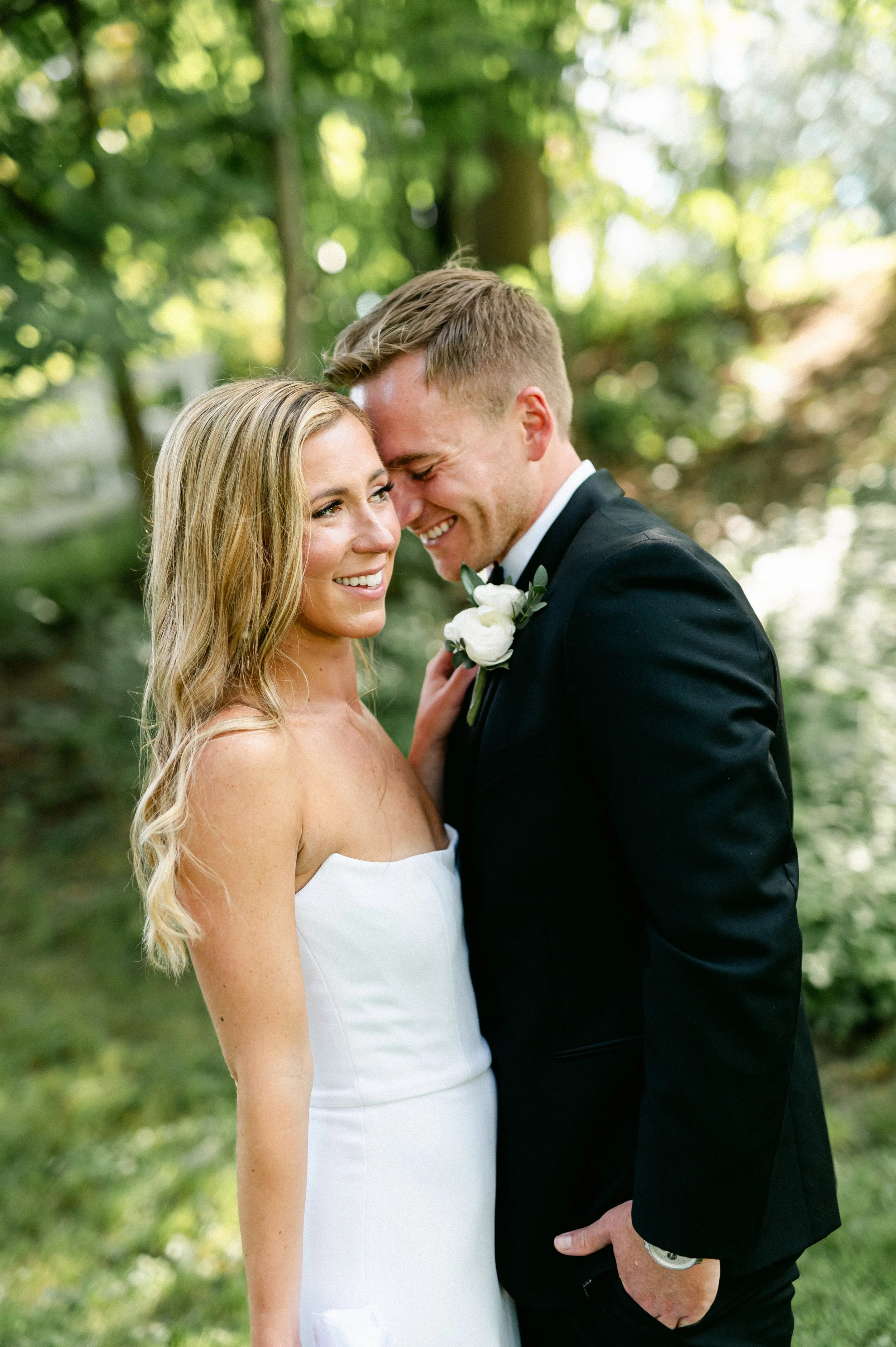 A newlywed couple stands close together outdoors in a wooded area, smiling with their foreheads touching. The woman has long blonde hair and is wearing a strapless white wedding dress. The man has short hair, wears a black tuxedo with a white shirt a