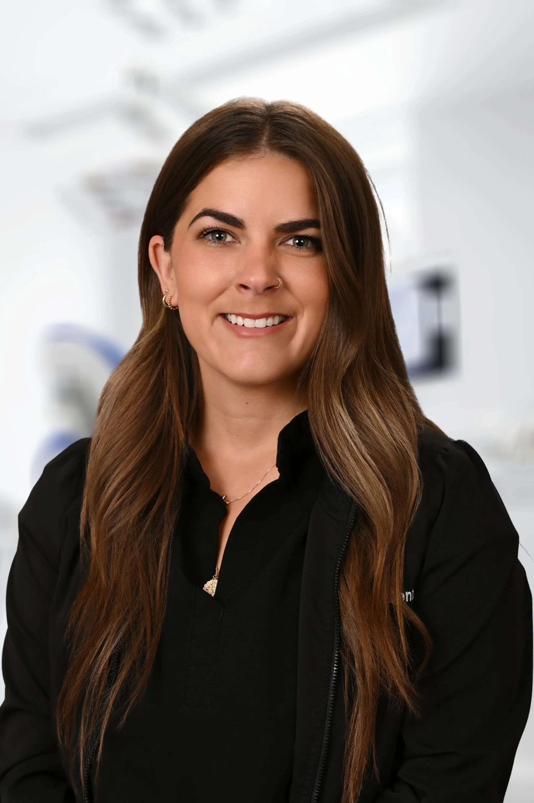 A smiling woman with long brown hair and a nose piercing, wearing a black jacket that reads 'Higgs Family Dental', standing in a dental clinic.