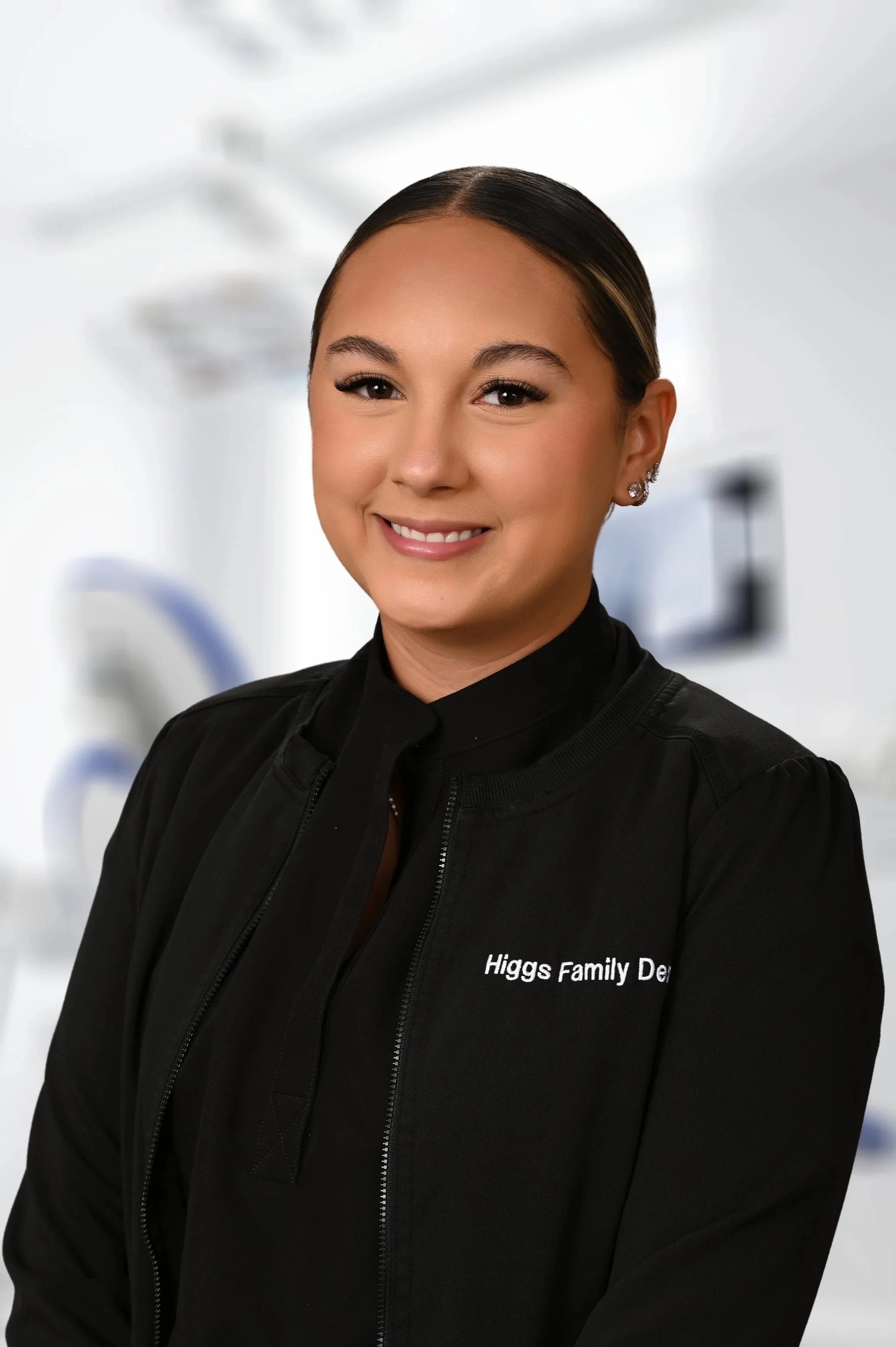 Dental professional wearing a black jacket with 'Higgs Family Dental' on it, smiling in a dental clinic.