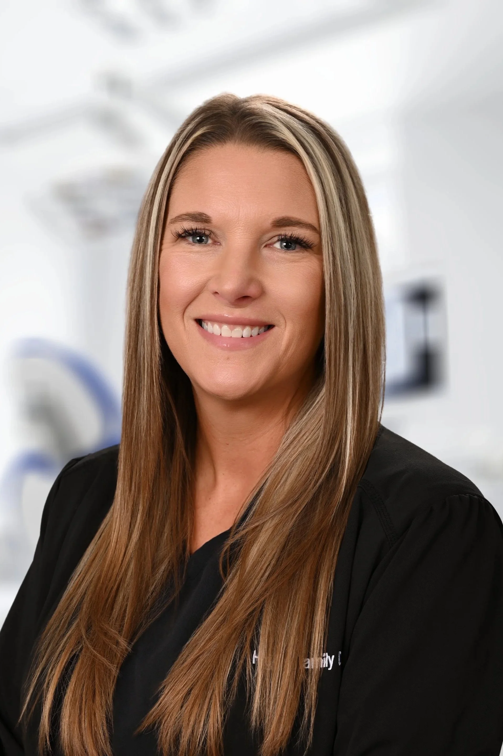 A smiling woman with long blond hair, wearing a black jacket with 'Family Dental' embroidered on it, in a dentist's office with dental equipment in the background.
