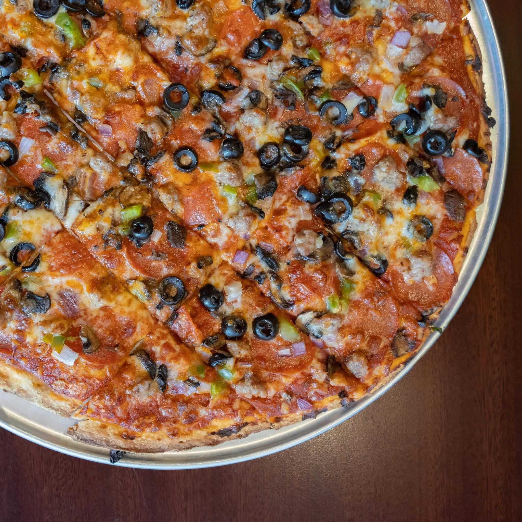 A close-up of a pizza in a silver pan on a dark wooden surface, topped with black olives, chopped onions, green peppers, and melted cheese.