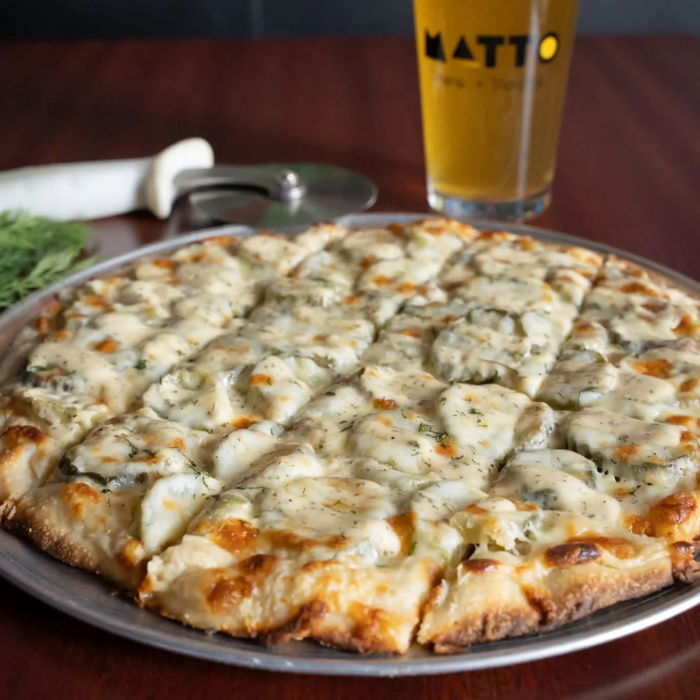 A cheese pizza with a golden crust on a round baking tray, with a glass of beer and pizza cutter in the background.