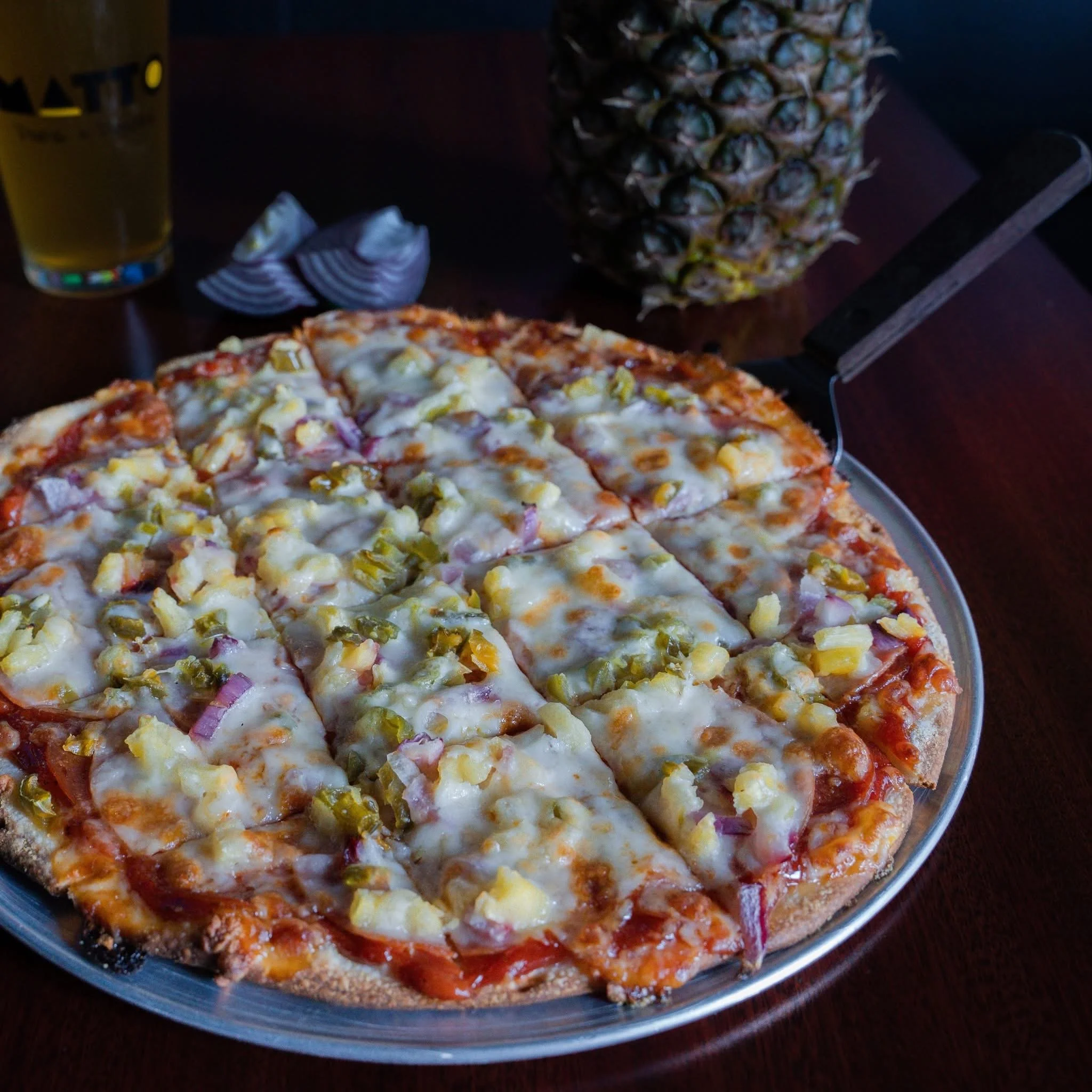A pizza topped with pineapple, red onions, and cheese, in a metal tray. In the background, a whole pineapple, a shell of red onion, and a glass of beer are visible on a dark wooden table.