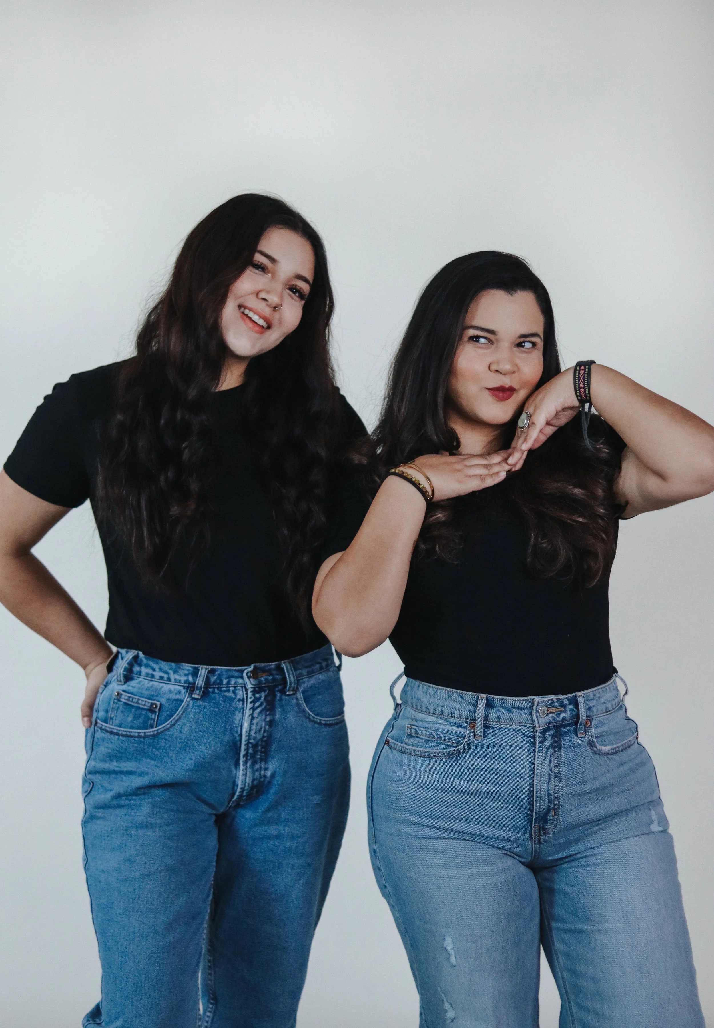 Two women with long dark hair wearing black t-shirts and blue jeans against a plain white background, posing playfully.