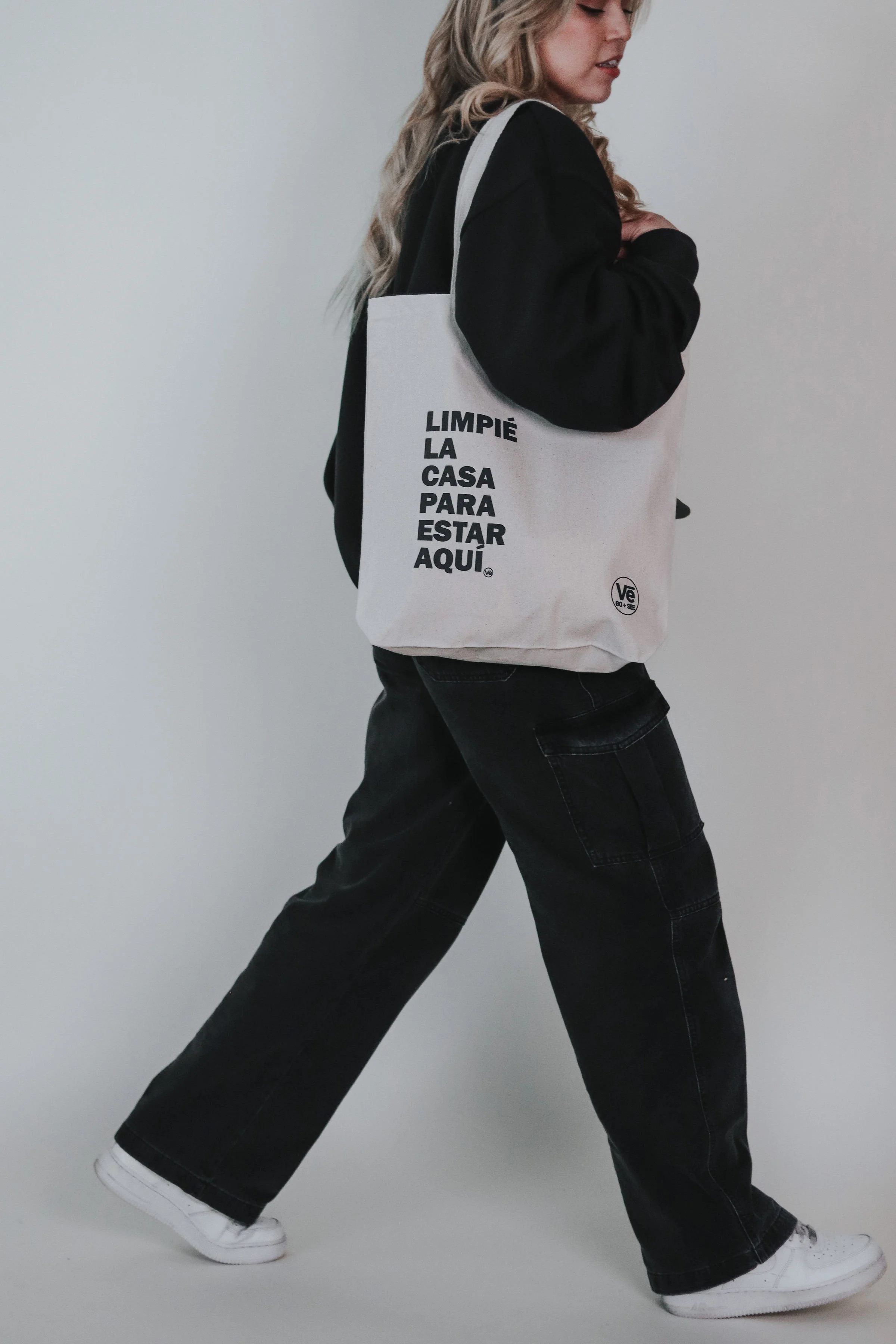 A person walking with a large off-white tote bag that has black text in Spanish, wearing black jeans, white sneakers, and a black top.