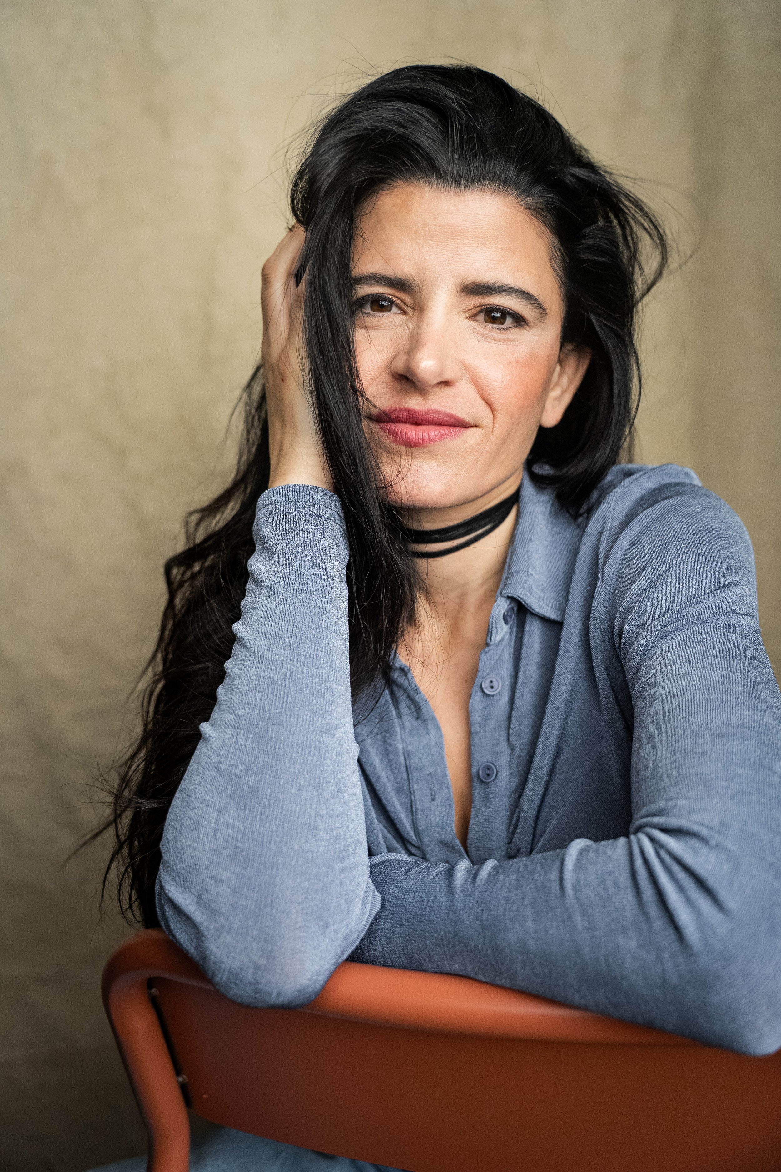 A woman with long black hair and light skin, wearing a light blue button-up shirt and a black choker, sitting in a terracotta chair with a beige background. She is smiling slightly and touching her hair with her right hand.