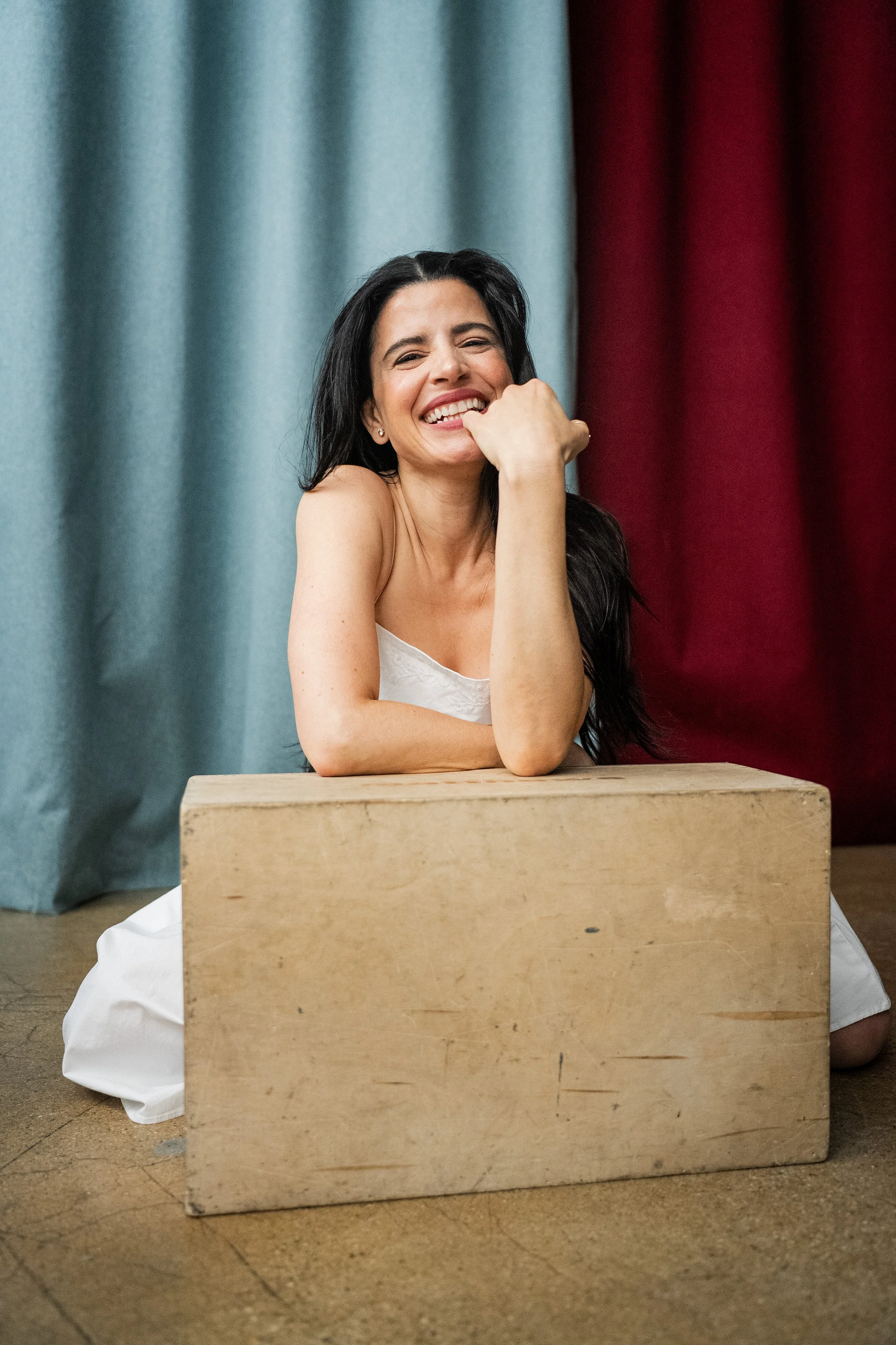 A woman with long dark hair smiling and resting her chin on her hand, sitting behind a large wooden box with curtains in blue and red colors in the background.