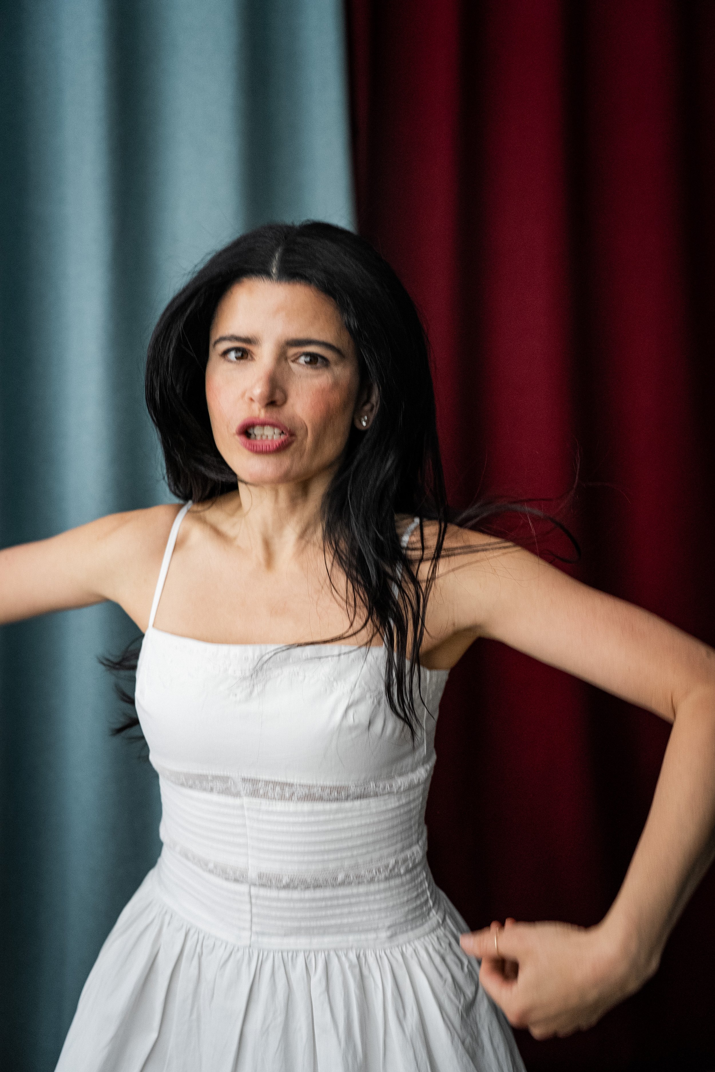 A woman with long dark hair and fair skin wearing a white dress with thin straps, standing with one arm bent at her waist and the other resting on her hip, in front of curtains that are teal on the left and red on the right.