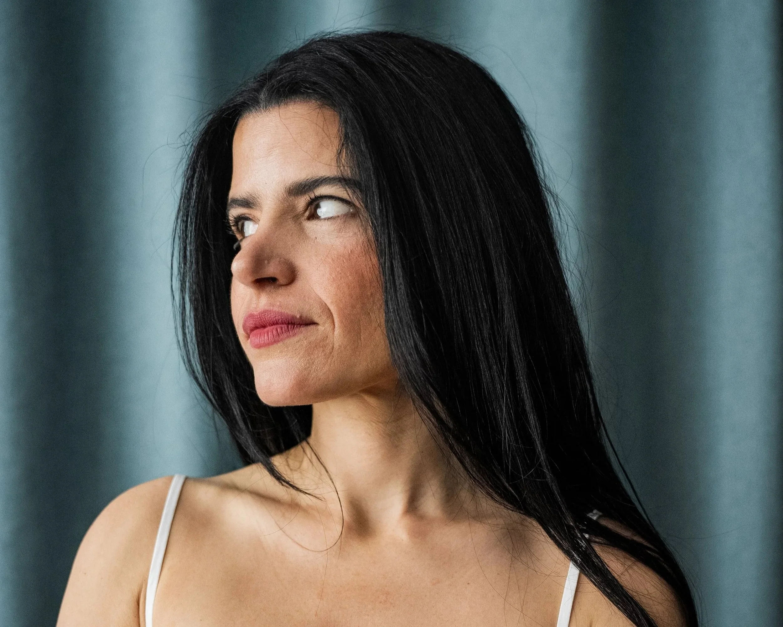 Close-up portrait of a woman with long black hair, tan skin, and wearing a white spaghetti strap top, looking slightly to her right against a teal background.