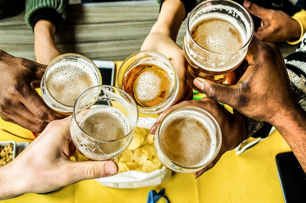 Six people clinking beer glasses together over a table with snacks.