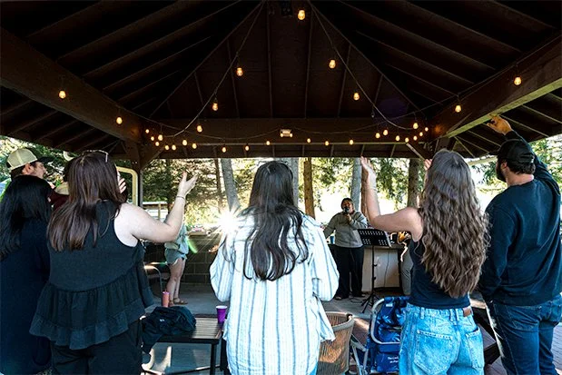 People singing and dancing on an outdoor covered stage with string lights, backed by trees.