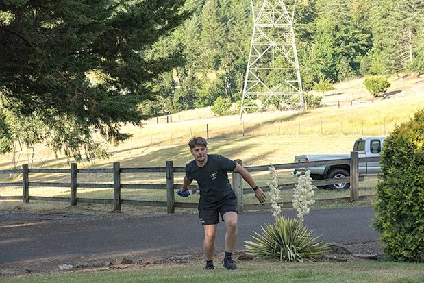 A young man in black shorts and a T-shirt looks surprised while running on the grass near a driveway with a wooden fence, a flowering plant, a parked white pickup truck, and a green forested hillside in the background.