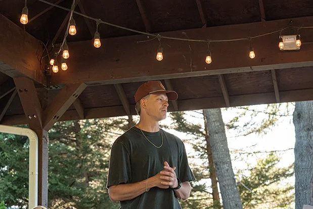 A young man wearing a brown cap, black T-shirt, and gold chain, standing outdoors under a wooden pavilion with string lights hanging overhead, in front of a background of trees.