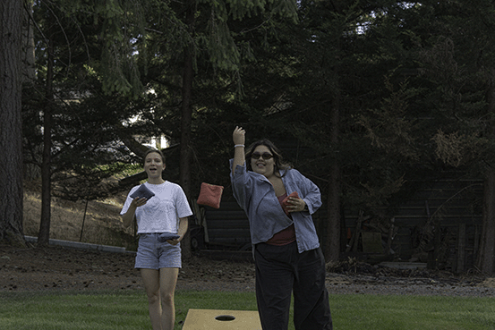 Two women playing cornhole outdoors on a grassy lawn surrounded by trees.
