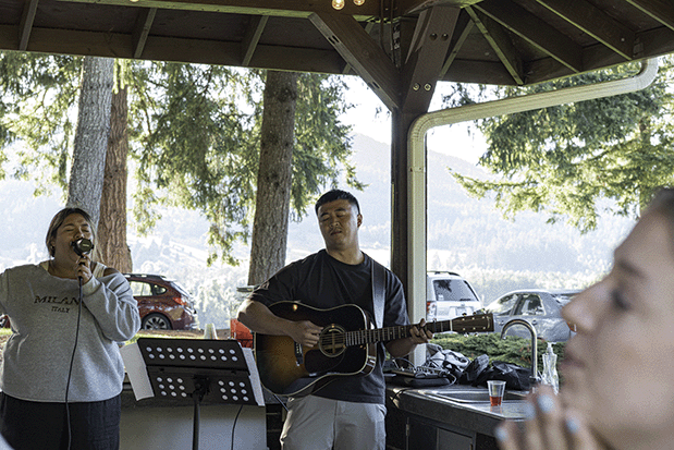 Two people singing and playing guitar under a wooden pavilion with trees and parked cars in the background.