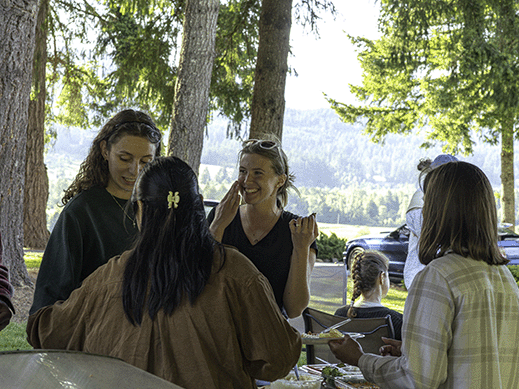 Group of people smiling and chatting outdoors near trees, with a lake and mountains in the background.
