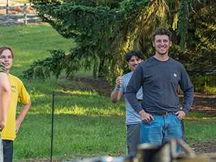 Three young men standing outdoors in a grassy area with trees, smiling and enjoying a sunny day.