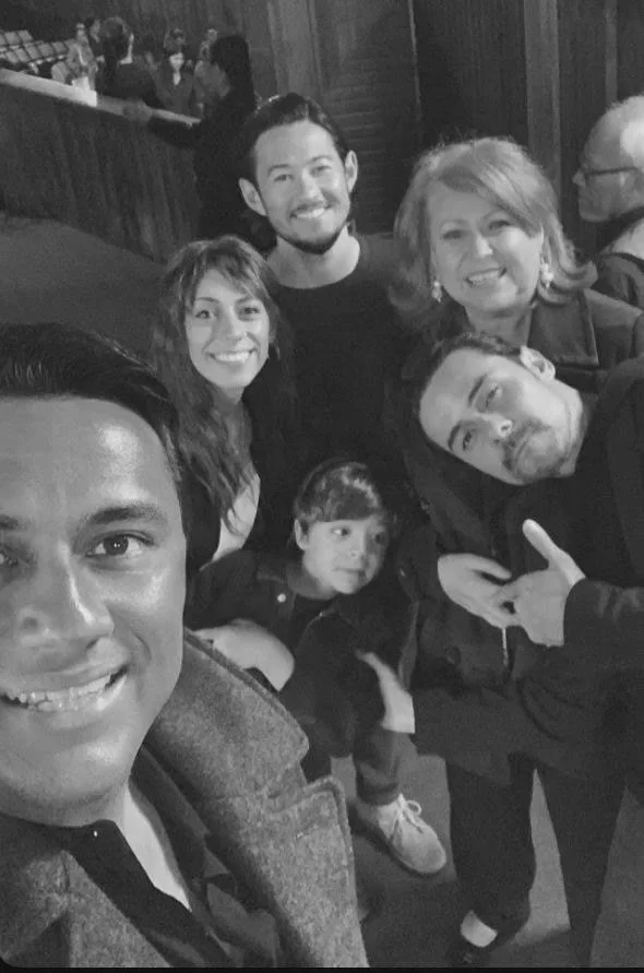 Group of six people taking a selfie at a social event or bar, smiling, with a bookshelf and other people in the background.