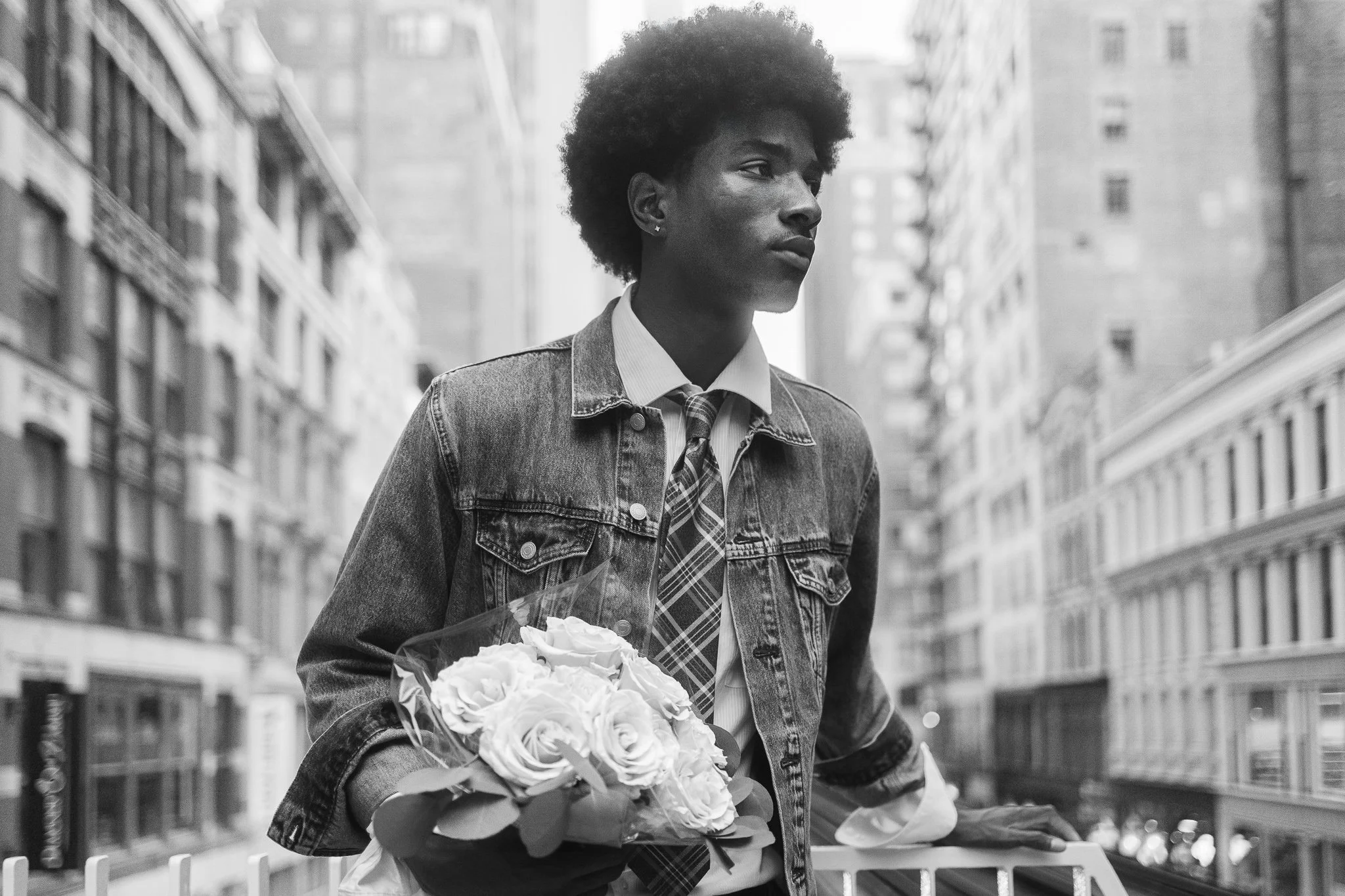 A black and white photo of a young man with an afro hairstyle standing on a city street, holding a bouquet of flowers at the train station