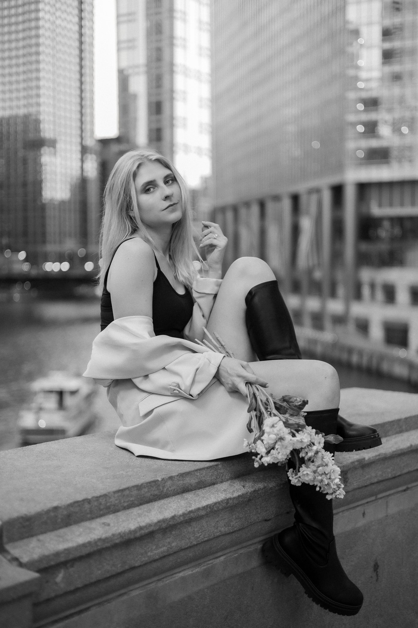 A black-and-white photo of a young woman sitting on a ledge in an urban setting. She has long blond hair and is wearing a sleeveless top, a light jacket, and tall black boots. A bouquet of flowers is placed in front of her.