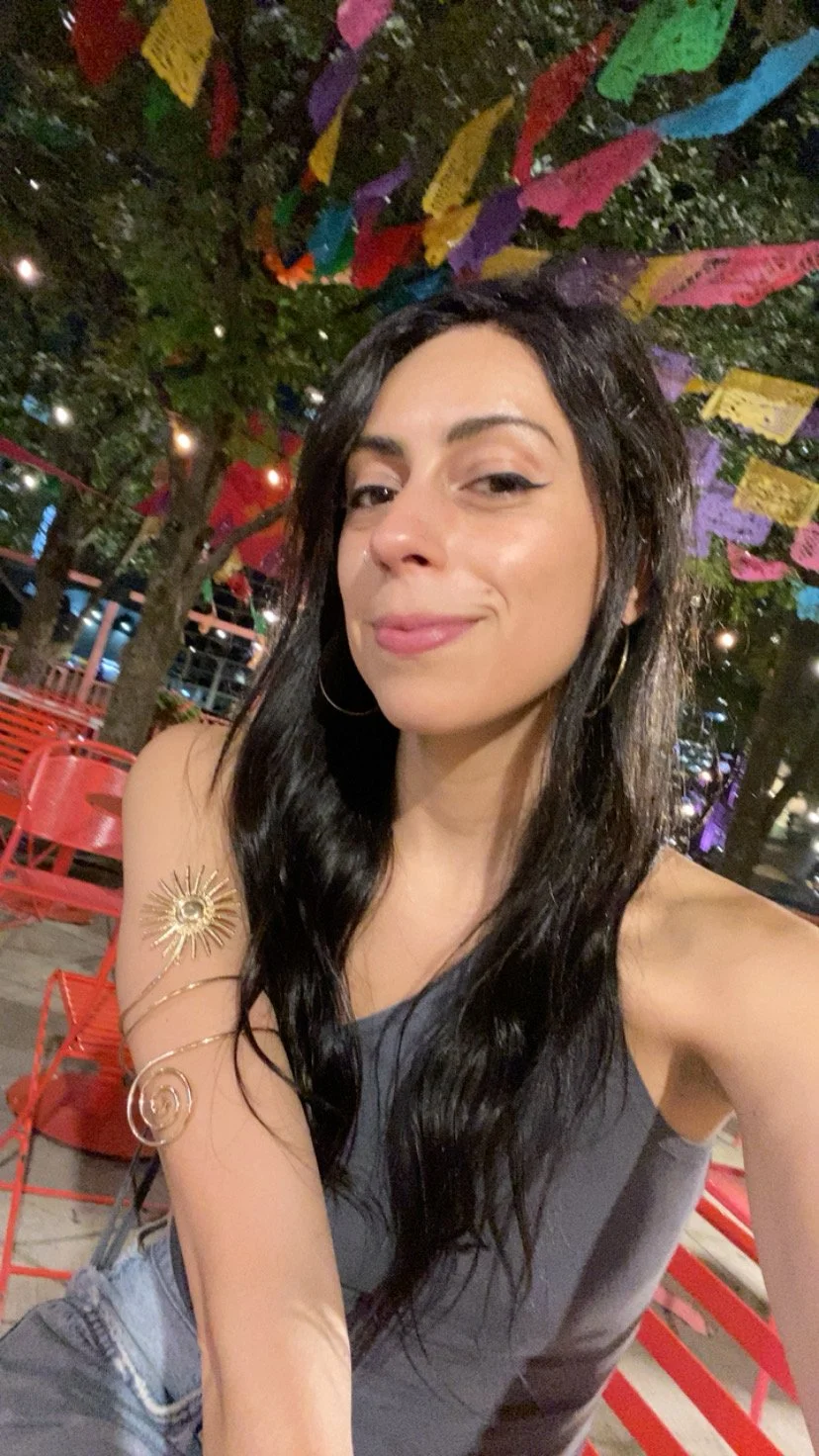 A woman with long dark hair and hoop earrings taking a selfie at night. She is wearing a sleeveless top with a decorative sun necklace on her shoulder. Behind her, colorful papel picado banners hang from trees, and red chairs are visible in an outdoo